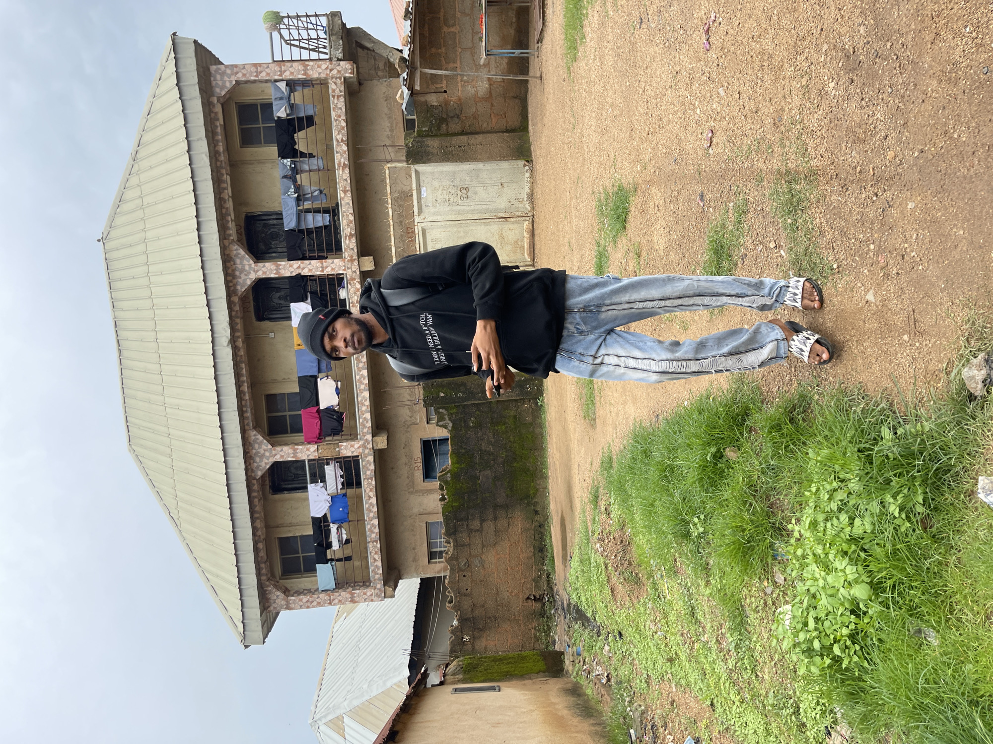 A young Black man is standing outside a multi-story building in Jos, Nigeria. He is wearing a black hooded sweatshirt, light blue distressed jeans, and white patterned slides. The building appears to be residential, with laundry hanging from the balconies. The ground is a mix of dirt and grass. The sky is overcast, suggesting it might be a cloudy day. The man is looking towards the camera with a neutral expression. The scene appears to be casual, possibly a street photograph. Text on the man's sweatshirt is partially visible and appears to read "DON'T NEED A BILLIONAIRE...". The overall impression is of an everyday scene in an urban environment in Nigeria.