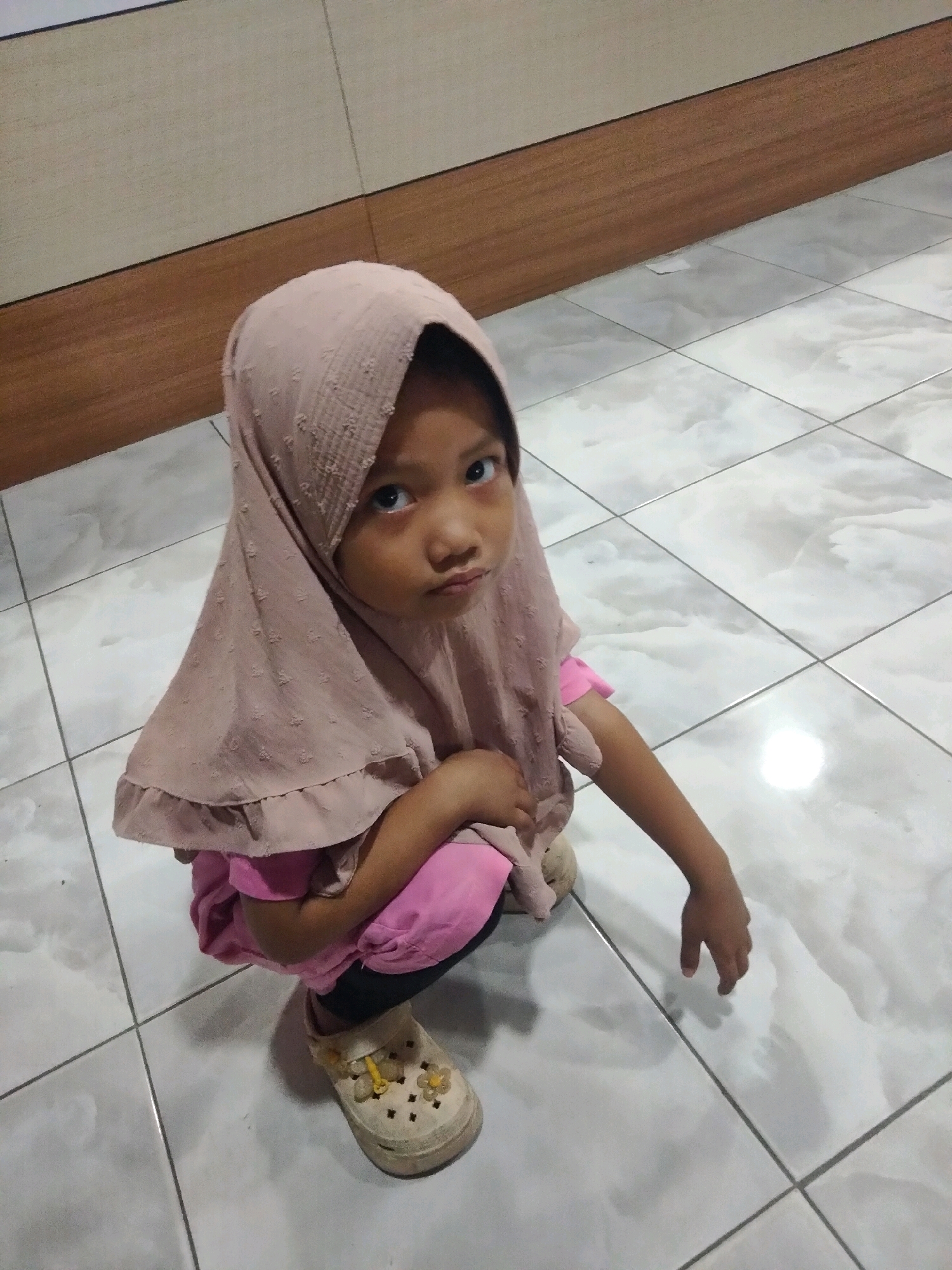 A young girl is crouching on a tiled floor. She is wearing a light pink hijab, a pink shirt, black pants, and light beige slip-on shoes with yellow accents. The floor is made of square, white tiles with grey grout lines. The background consists of a wall with a beige upper section and a darker wooden paneling lower section. The lighting suggests an indoor environment.
