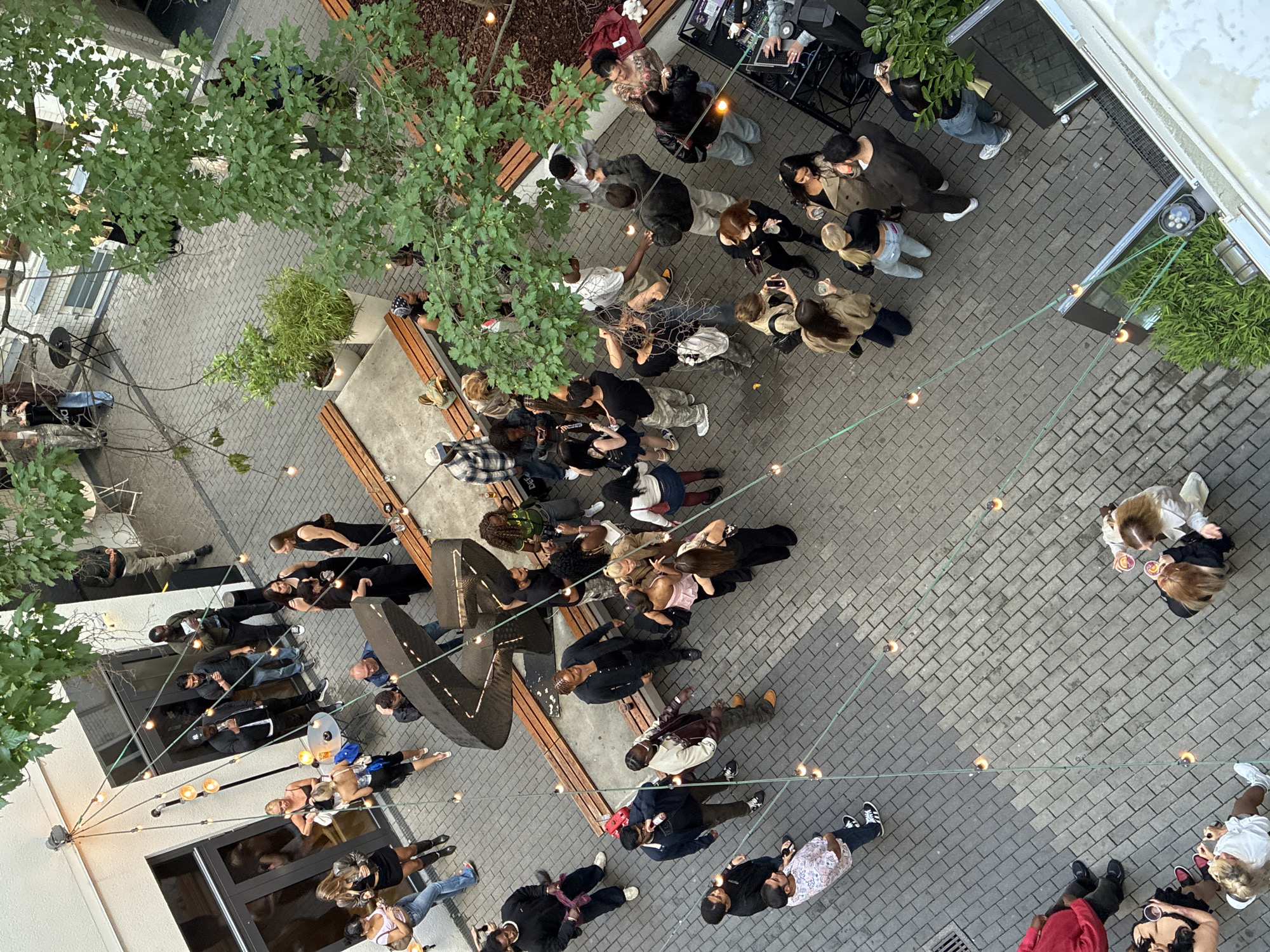 This image captures a bustling outdoor courtyard event, likely at a venue in Berlin, Germany, suggested by the modern, urban architecture and the style of the people. The scene is densely populated with individuals engaged in various social activities.
In the foreground, a group of women are gathered, holding drinks and interacting. To the left, a man in a red shirt is seated, and another person in a white top is standing nearby, also with a drink. Further to the right, a DJ setup is visible with a person operating the equipment. Surrounding the courtyard are buildings with large windows and doors, and interspersed greenery adds to the ambiance. String lights are draped across the space, casting a warm glow on the paved ground.
The crowd consists of people of diverse ethnicities and ages, predominantly dressed in casual to semi-formal attire. Many are holding drinks, conversing, or observing the surroundings. Some individuals are standing in small groups, while others are more isolated. The overall atmosphere appears to be one of a social gathering, possibly a party or an outdoor bar setting. The perspective is from an elevated viewpoint, looking down into the courtyard.