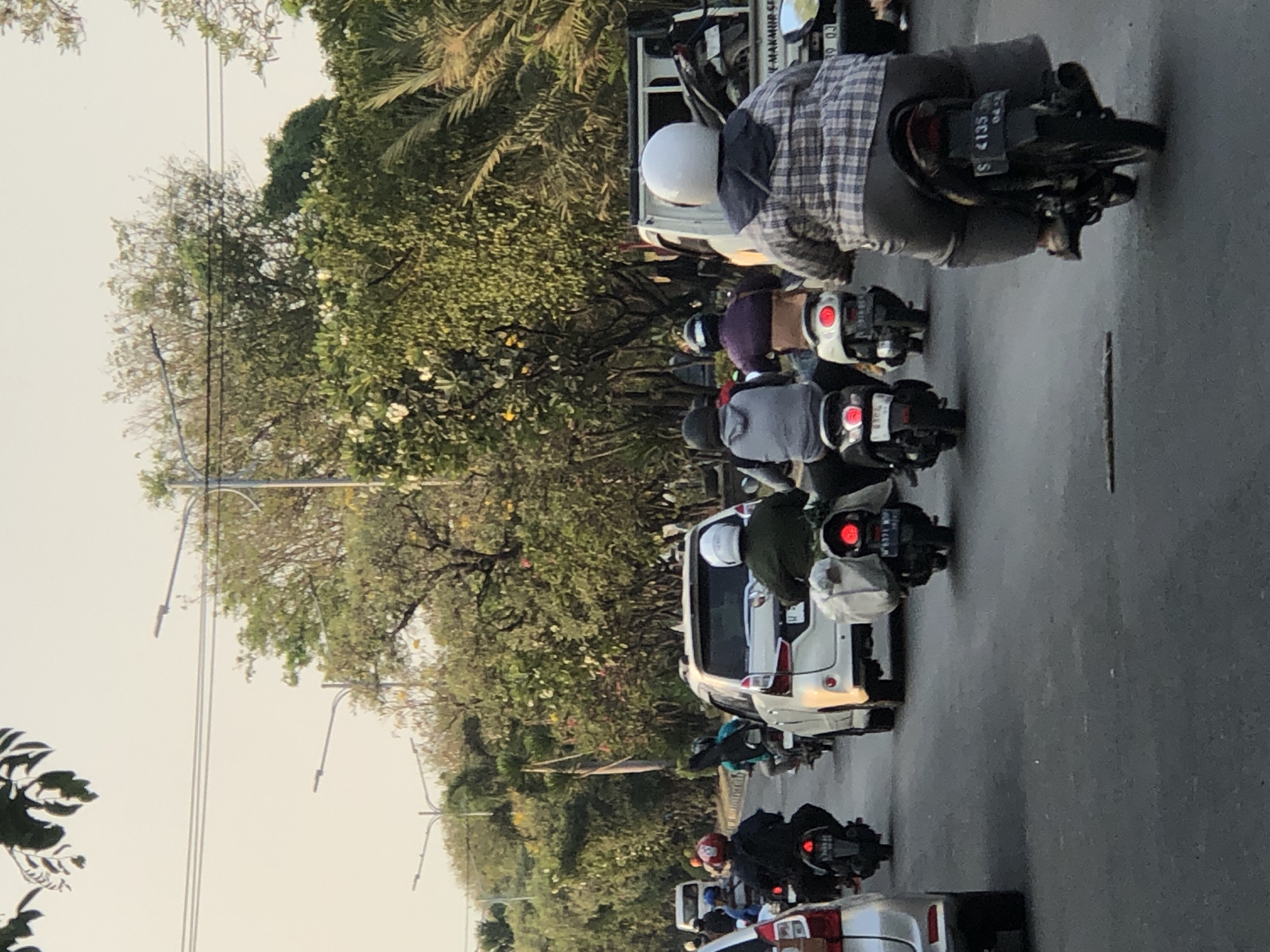 In a city street scene in Ngaban, Indonesia, a moderate flow of traffic is depicted. Several motorcycles, occupied by riders wearing helmets and casual attire, are on the road. One rider is wearing a plaid shirt, and another is in a dark green jacket. A white car is in the middle lane, and a light-colored pickup truck is to its right. Trees line the street, and streetlights are visible overhead. The sky is overcast, suggesting it is either early morning or late afternoon.