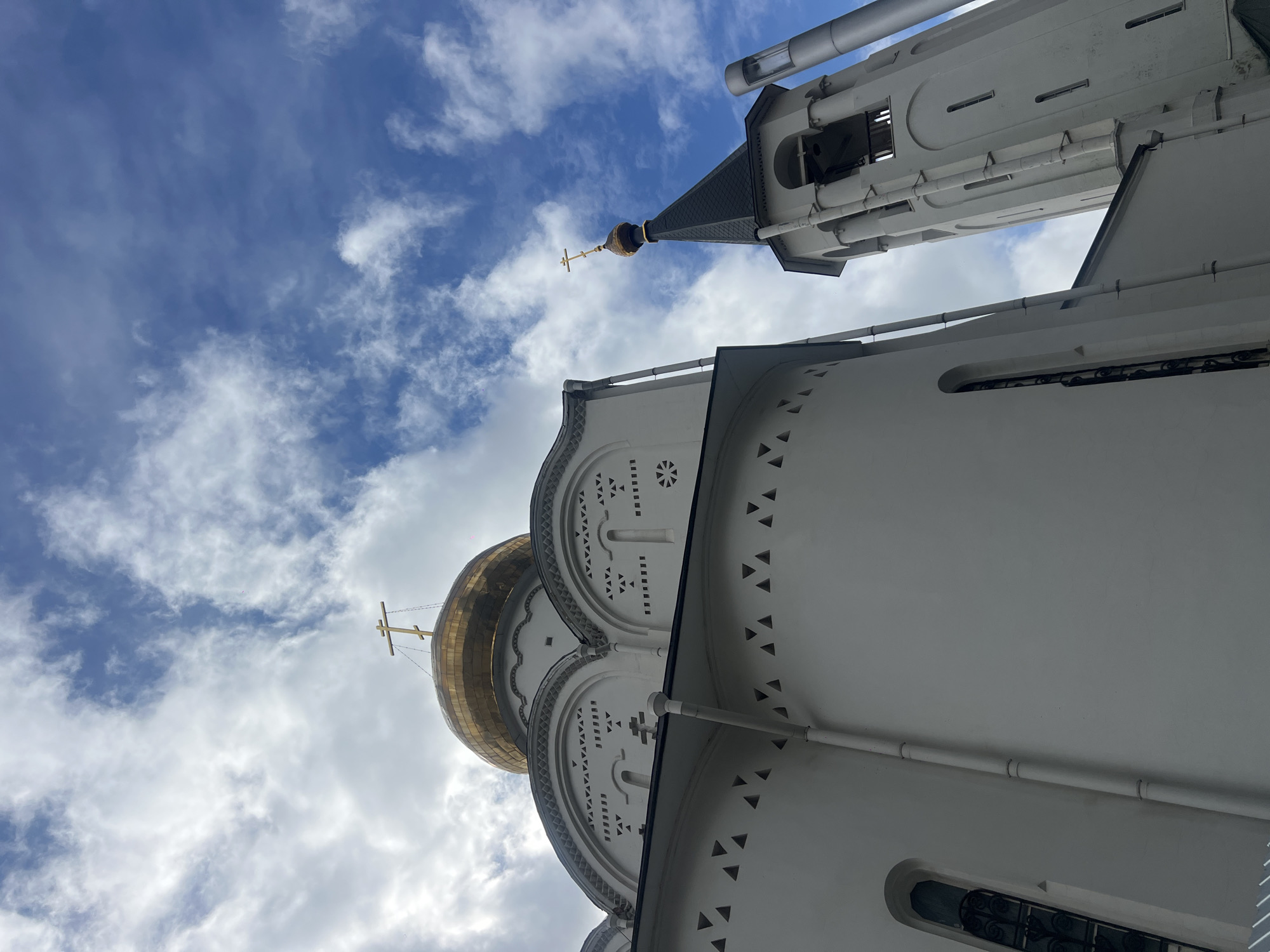 An upward shot showcases architectural features of a building complex under a partially cloudy sky. The complex includes a golden, onion-shaped dome with a cross on top, typical of Russian Orthodox churches, and a white tower with a spire and another cross. The buildings are white with decorative architectural details. Based on the architectural style, the image likely depicts a structure within Moscow, Russia. No people are visible in the frame.