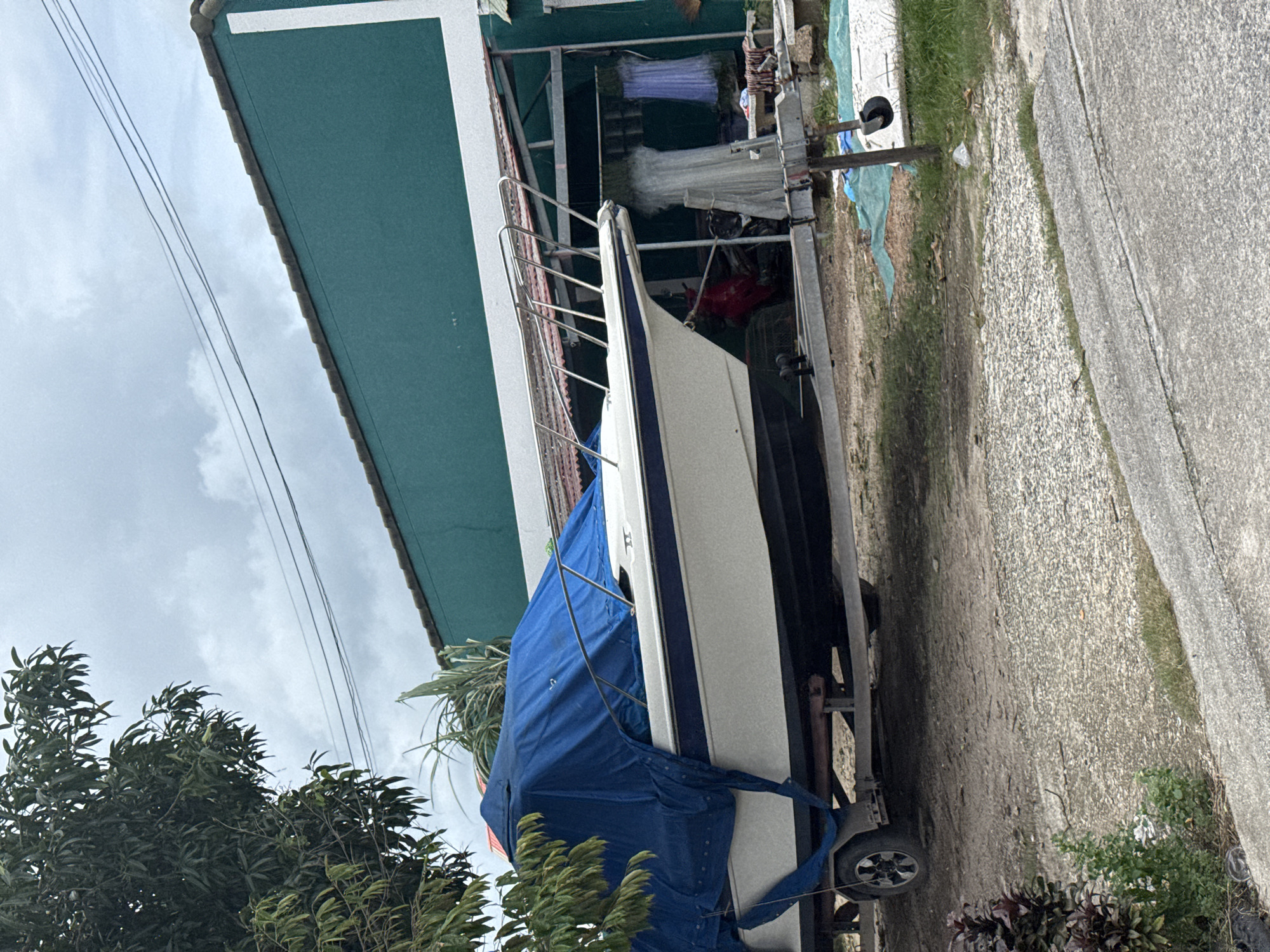In Rawai, Thailand, a white and blue boat, partially covered by a blue tarp, rests on a trailer in a dirt lot. The boat is positioned in front of a green building with a corrugated roof, which appears to be a residence or business. To the left of the boat, a large tree with dark green leaves is visible. In the background, a cloudy sky looms overhead, with power lines stretched across it. The foreground features a concrete road and some patches of grass. The scene appears to be one of storage or temporary parking for the boat.