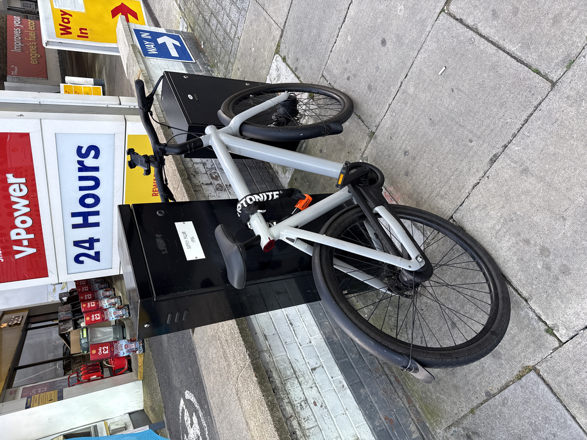 A light grey VanMoof bicycle is locked to a black supply pillar on a sidewalk in London, United Kingdom. The pillar is adjacent to a Shell gas station, with visible signage indicating "V-Power 24 Hours" and "Way In." Promotional materials for the gas station, including "Only £2," are visible. The surrounding environment consists of concrete sidewalk and street-level details typical of an urban setting. The bicycle's lock is an orange Kryptonite.