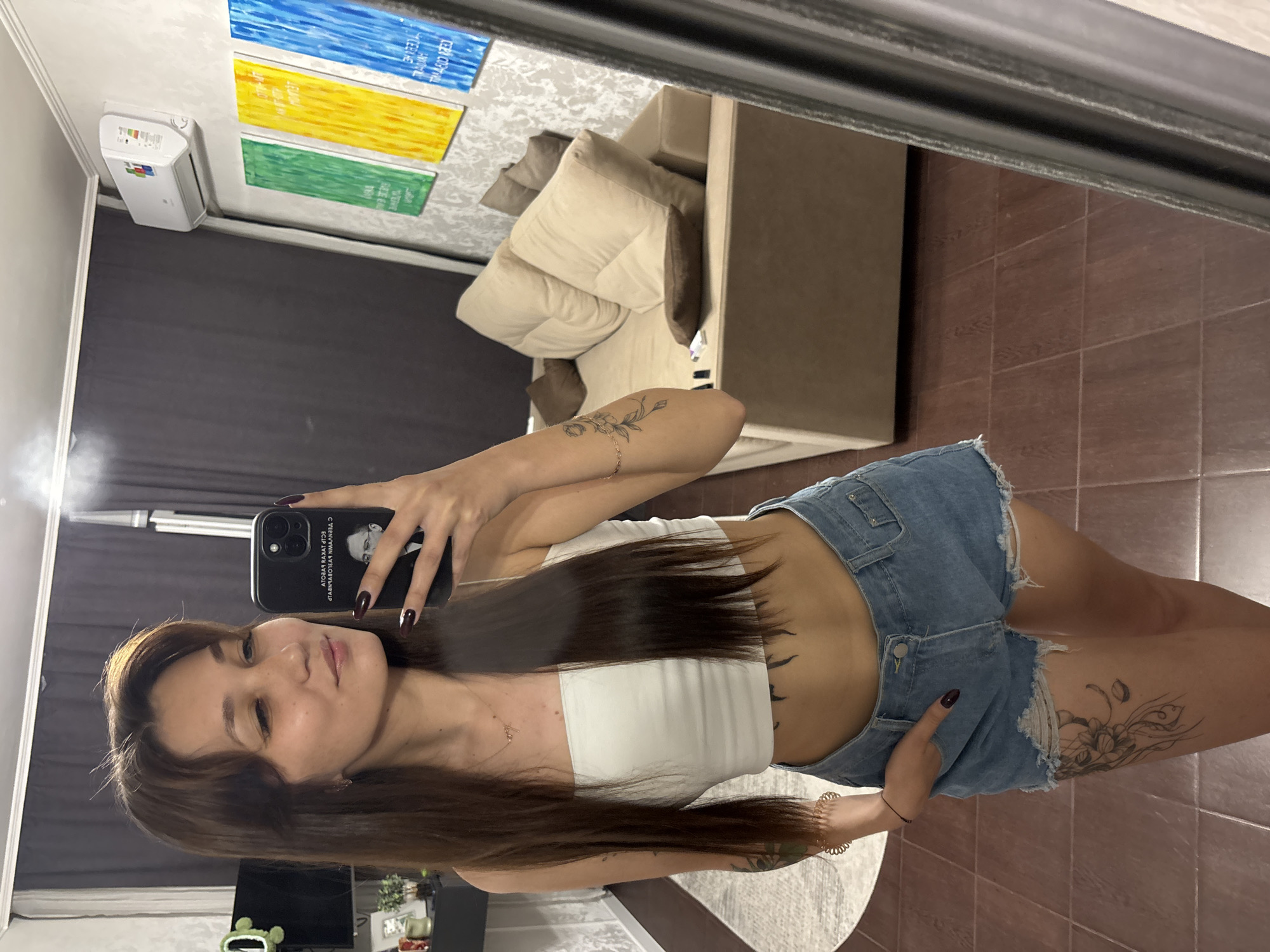 A young woman with long brown hair is taking a mirror selfie in what appears to be a home interior. She is wearing a white bandeau top and denim shorts, with visible tattoos on her arms and legs. She is holding a smartphone in her right hand, which is angled to capture the reflection of herself and the room.
The room is furnished with a large, light-colored sofa with several cushions. Behind the sofa, there are three colorful abstract paintings on a wall. An air conditioning unit is mounted on the wall above the paintings. The floor is tiled in a dark reddish-brown color. The overall impression is of a casually decorated living space.
While the image provides details of the woman and her immediate surroundings, there is no information within the image itself to determine the specific location in the city of Novokuznetsk, Russia.