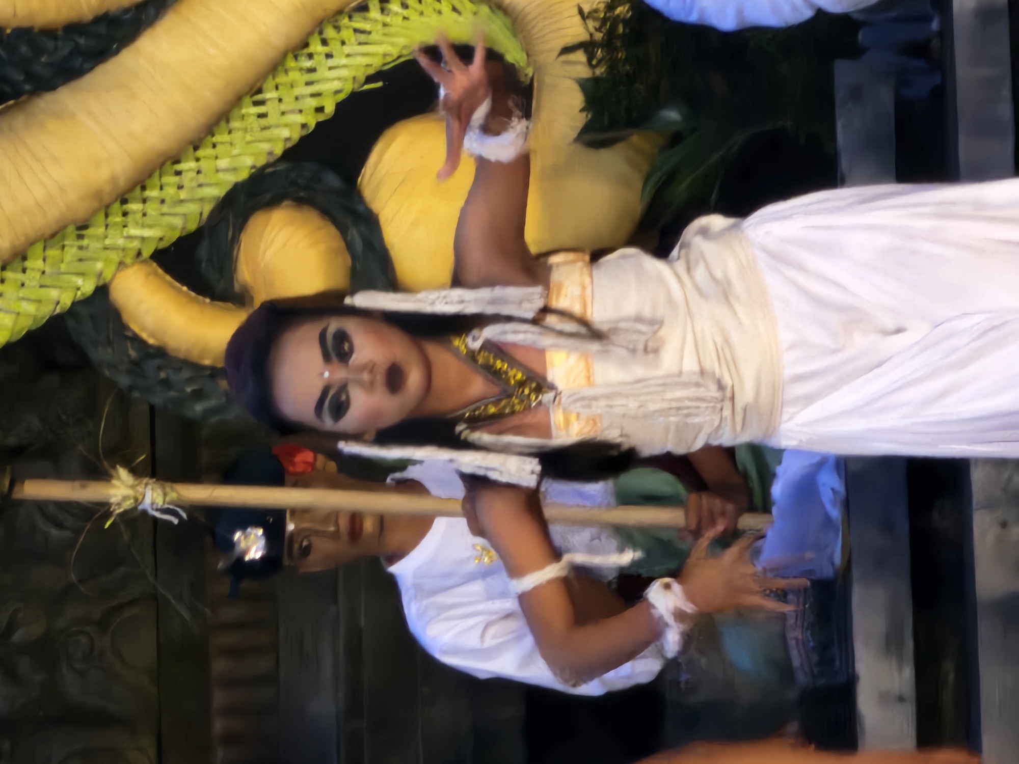 A staged performance is taking place in Denpasar, Indonesia. Two individuals, one in the foreground and another slightly behind and to the left, are dressed in traditional attire. The individual in the foreground is a female dancer adorned with heavy makeup, including dark eyeliner and lipstick, and wears a white costume with ornate gold trim around the neckline. She has white fabric strips hanging from her head and is wearing white bracelets on her wrists. Her hands are outstretched, palms facing forward, in a graceful pose. She is standing near a large, coiled object that appears to be a decorative representation of a snake, with yellow and green woven textures. The second individual, positioned behind the dancer and holding a wooden staff, is dressed in a white long-sleeved shirt and wears a dark headdress with a red accent and a small jeweled emblem. The background is dark and appears to be an outdoor setting, possibly a temple or performance venue, with blurred wooden steps visible.