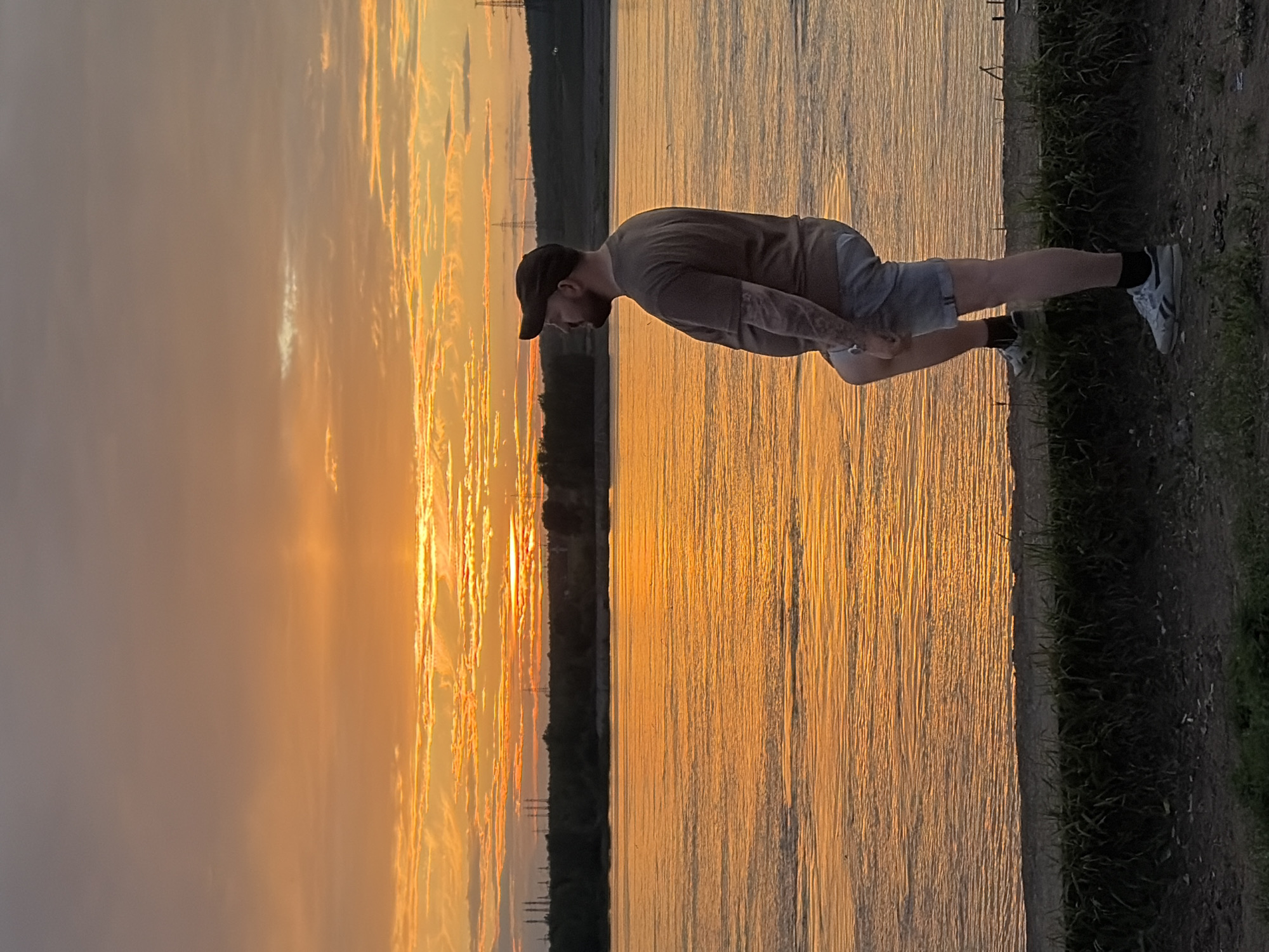 The media file shows a man performing a plank exercise next to a body of water. The man is wearing a grey t-shirt, grey shorts, a black baseball cap, black socks, and white sneakers. He has tattoos on his left arm. The setting is outdoors, with a large expanse of water reflecting the sunset. The sky is filled with dramatic clouds illuminated by the setting sun, casting a warm orange glow over the scene. The water is calm with ripples, and the shoreline is visible with grass and dirt. The time of day is sunset, indicated by the warm lighting and colorful sky. The location is Chaikovsky, Russia. The man appears focused and engaged in his exercise. There is no visible text in the image.