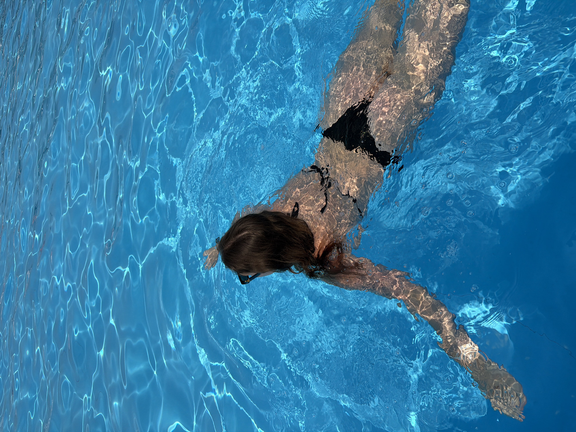 A person is swimming in a bright blue swimming pool. The person is wearing a black bikini and sunglasses. The water is clear and reflects sunlight, creating shimmering patterns on the surface. The person is seen from behind, with their arm extended in front of them, propelling them through the water. The setting appears to be an outdoor pool. The location is specified as Drofkino, Russia, suggesting a public or private swimming facility within that area.