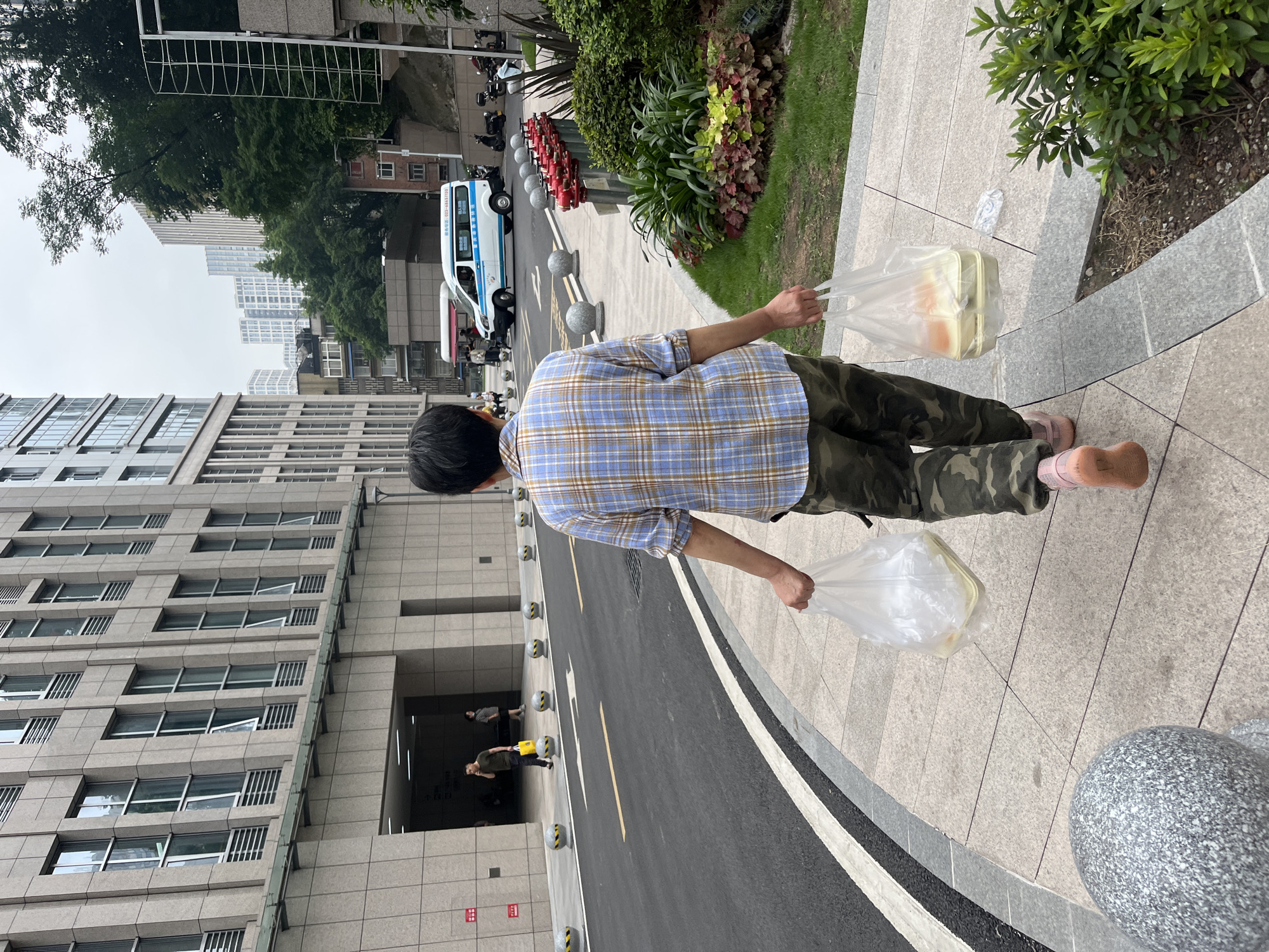 The image shows a person from the back, walking on a sidewalk in front of a modern building in Yuzhong District, China. The person is wearing a blue and brown plaid shirt, camouflage pants, and pink shoes. They are carrying two plastic bags, each containing food in a takeout container. To the left of the person is a street with a white and yellow striped line, and to the right is a grassy area with plants and some red fire hydrants. In the background, there are more buildings, trees, and a white van. The sky is overcast.