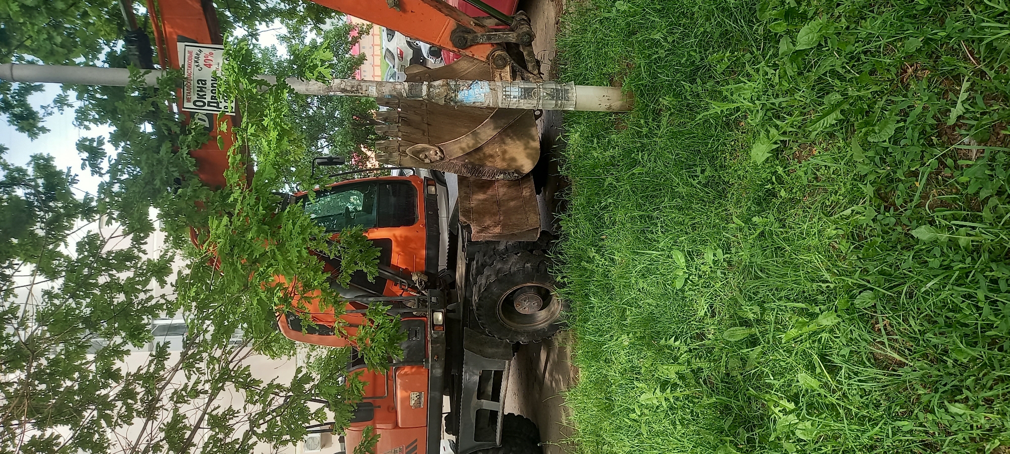 An orange backhoe rests on a paved surface abutting a grassy area. The backhoe's bucket is suspended near a slender pole covered with fliers. A sign advertising windows is affixed to the pole. Part of a building is visible in the background. The location is Krasnodar, Russia.