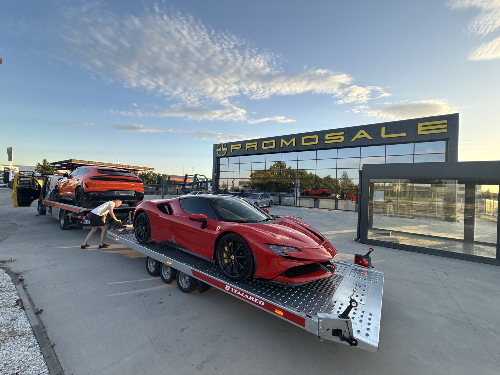 The image shows a red Ferrari SF90 Stradale being unloaded from a silver TEMAREd trailer. Another vehicle, a bright orange SUV (likely a Lamborghini Urus), is on the truck above. A man wearing a white shirt and black shorts is present, appearing to guide or assist in the unloading process. In the background is a modern building with the large sign "PROMOSALE" in yellow letters. Cars are reflected in the building's mirrored windows. The location appears to be an automotive dealership or showroom, likely within the city of Plovdiv, Bulgaria. The sky is blue with scattered clouds. The day is bright with some shadows visible.