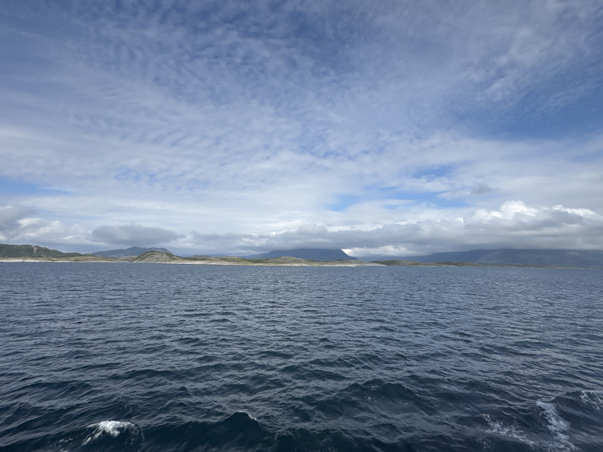 The image depicts a panoramic view of the ocean off the coast of Onøya, Norway. The foreground is dominated by the deep blue, undulating surface of the sea, with small white wave crests visible. In the midground, a low-lying, rocky coastline stretches horizontally across the frame, dotted with sparse vegetation. The land appears rugged and sparsely populated. The background features rolling hills and mountains, partly obscured by a layer of clouds. The sky is a mix of blue patches and white, wispy clouds, suggesting a dynamic weather system. The overall impression is one of a vast, open, and somewhat wild coastal landscape.