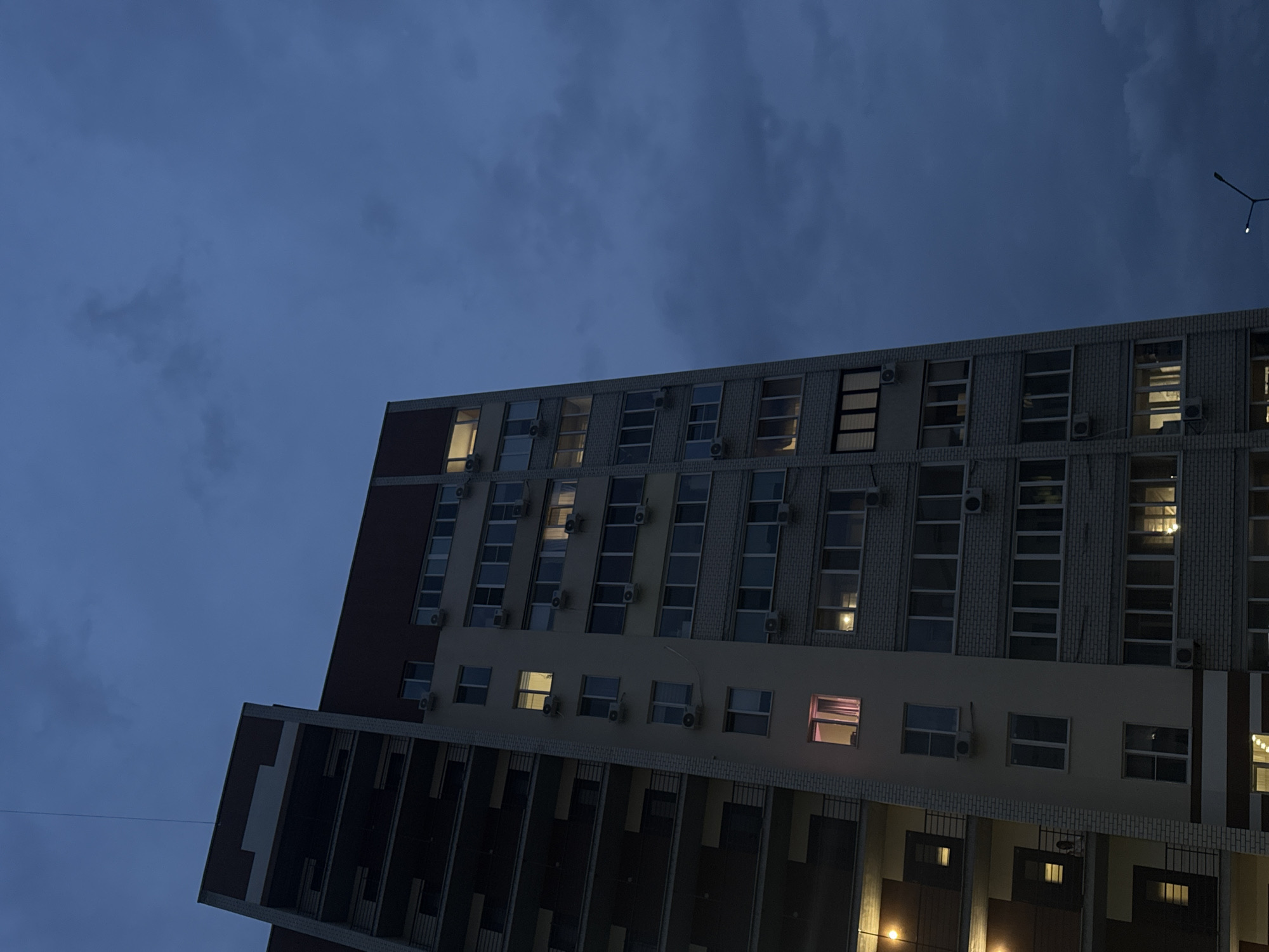 A low-angle shot of a high-rise building under a cloudy, dark blue sky at dusk or night. The building has a rectangular blocky design with a mix of dark brown and light beige sections. Each level has a row of windows, many of which have air conditioning units installed beneath them. Some windows are illuminated from the inside, revealing a warm, yellow glow while one has a distinct pink hue. A streetlight is visible in the upper right corner of the frame. This is located in the city of Saratov, Russia.