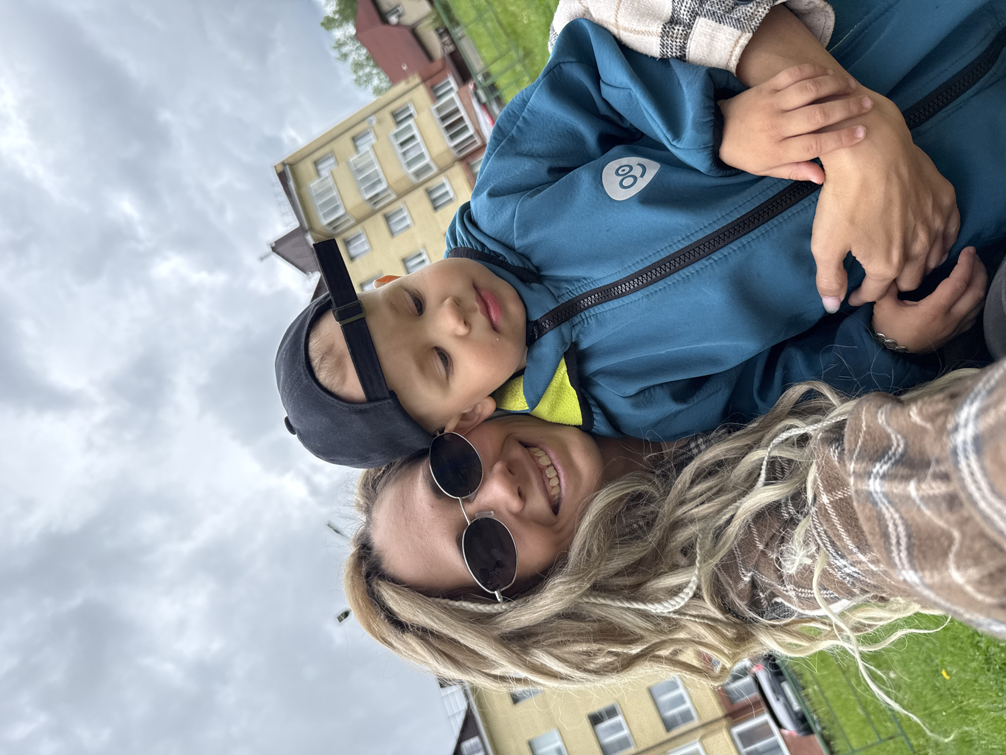 A young woman with long, blond dreadlocks and round, dark sunglasses is lying on her back in what appears to be a grassy area. She is smiling and looking directly at the camera. A young child is lying on top of her, resting on her chest, wearing a dark baseball cap and a blue jacket. The child's eyes are closed. The woman is wearing a brown and white checkered flannel shirt. In the background, there is a multi-story building in a light yellow color. The sky is overcast with grey clouds. This scene is at the location of Rusanova, Russia.