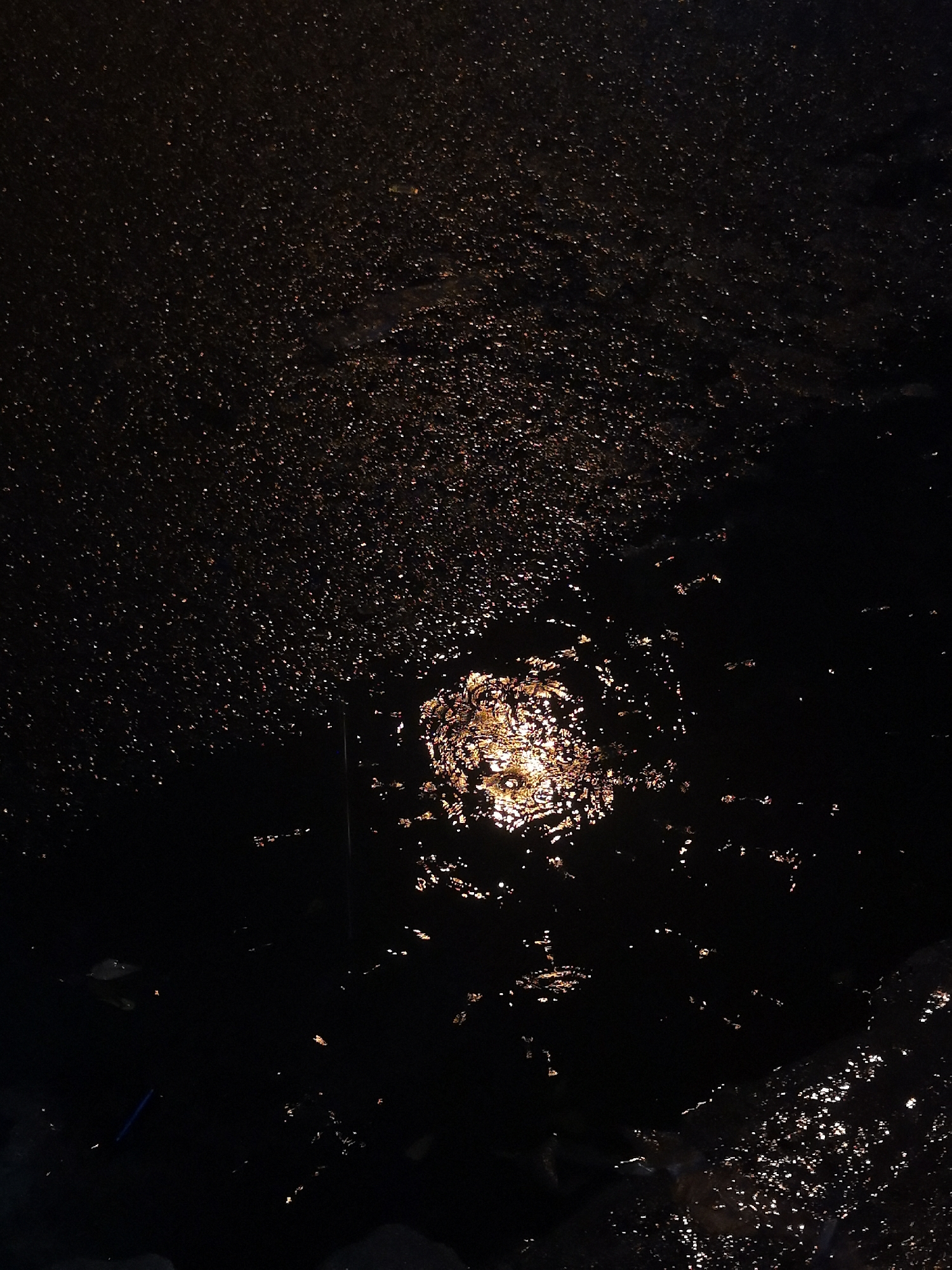 A low-angle, vertically-oriented image shows a dark, uneven surface covered in shiny reflective droplets, suggesting a wet or damp asphalt ground. In the central area of the image, a concentrated, bright reflection illuminates the surface, with a circular pattern resembling the light source. The remainder of the surface has a scattering of smaller, less intense reflections. In the bottom portion, some dark, larger shapes suggest stones. There's a small blue object in the bottom left. Without additional information, no specific location within Bac Nha Trang Ward, Vietnam, can be identified. There are no people or subjects in the image.