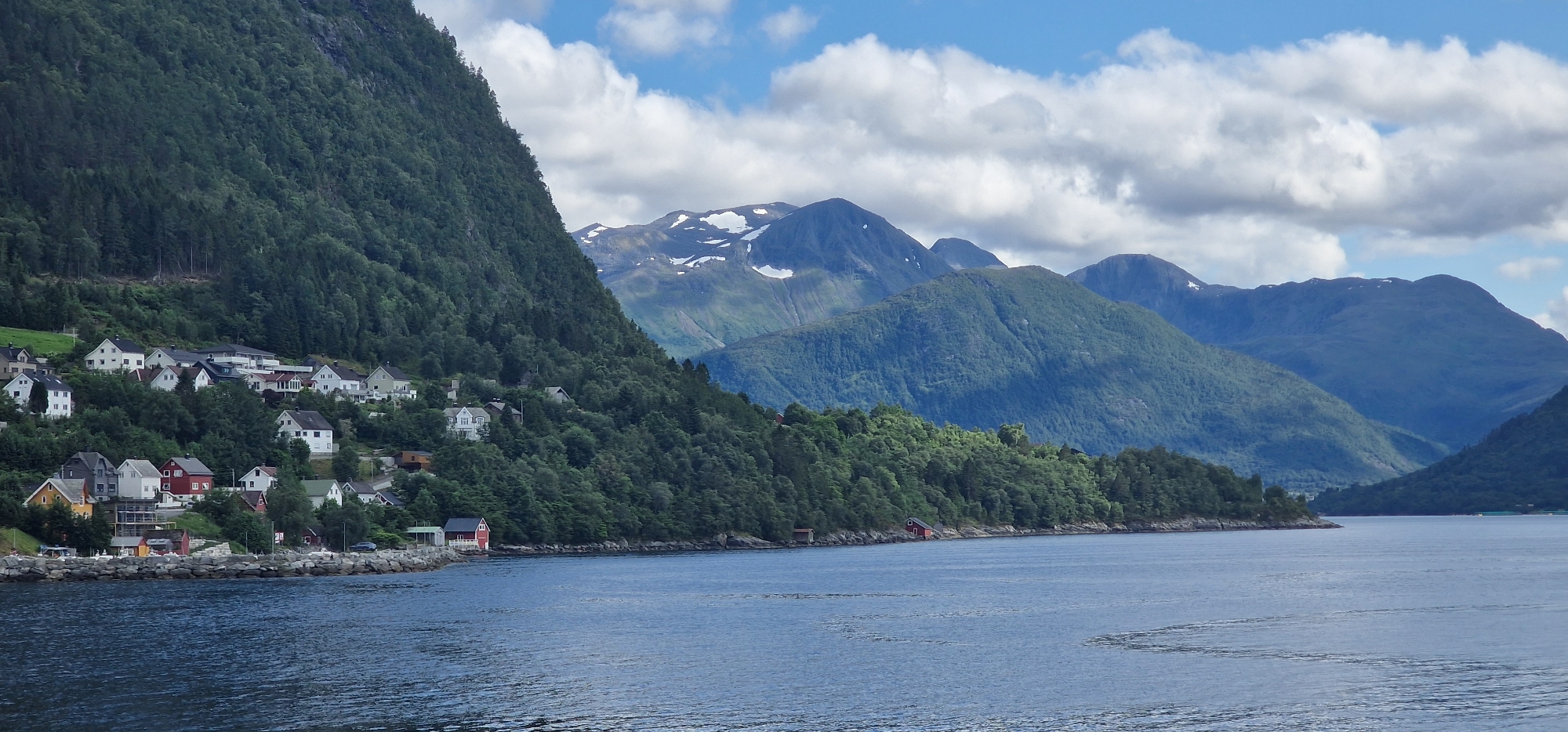 The image depicts a scenic view of a residential area nestled on a steep, tree-covered mountainside overlooking a body of water. The architecture consists primarily of white and light-colored houses with red roofs, typical of Norwegian villages. The settlement appears to be situated in the city of Volda, Norway, a location known for its dramatic fjords and mountainous terrain. The water in the foreground is calm, reflecting the cloudy sky and the surrounding landscape. In the background, imposing mountains rise, some with snow-capped peaks, indicating a high latitude or elevation. The overall impression is one of tranquility and natural beauty, characteristic of the Norwegian countryside. There are no visible people or animals in the scene, and the environment appears undisturbed.