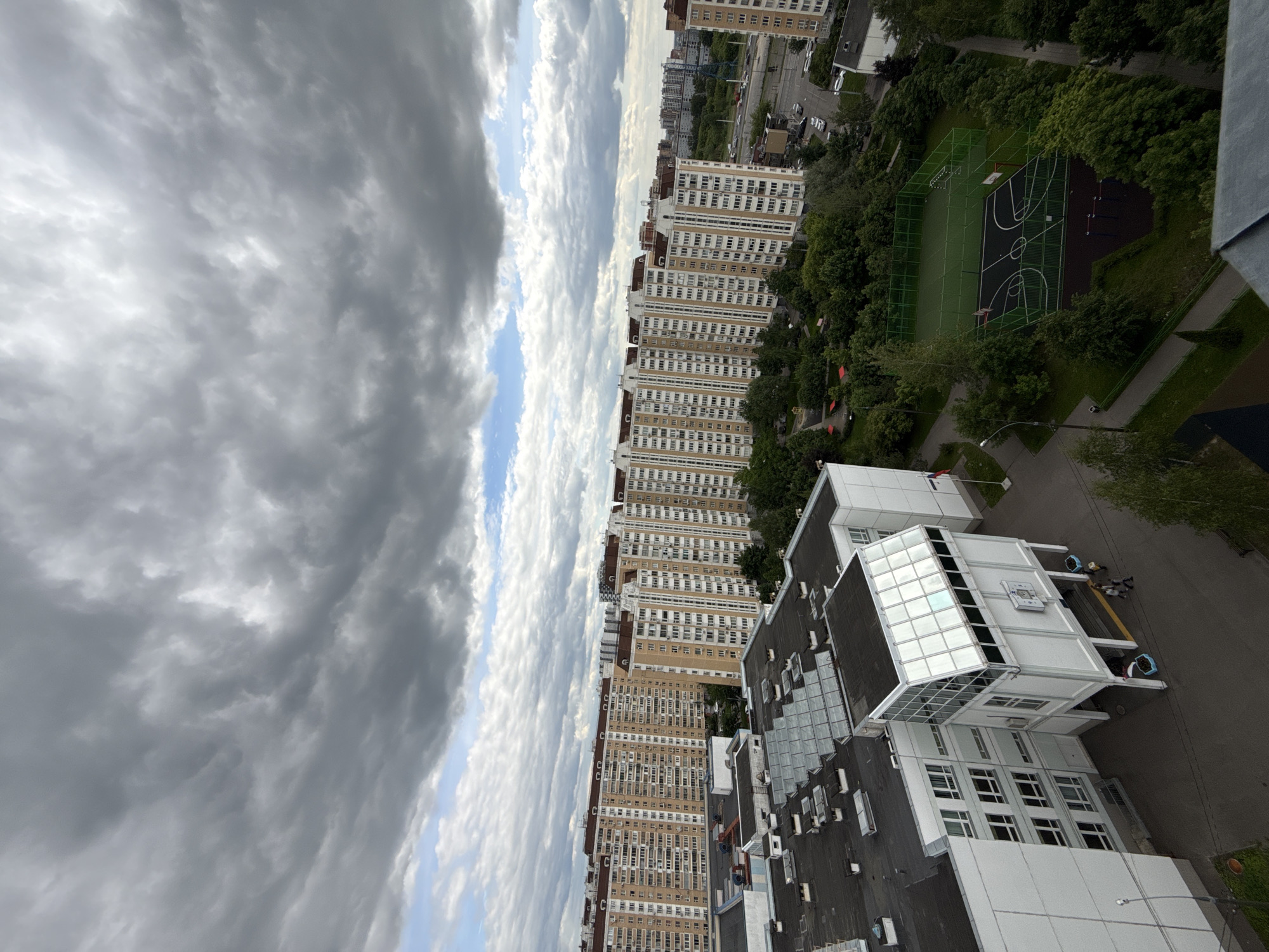 The scene depicts an urban environment in Moscow, Russia, viewed from an elevated position, possibly a high-rise building. The image shows a cluster of tall, multi-story residential buildings extending towards the horizon, with a clear division between a densely clouded sky and the urban landscape. Below the apartment buildings are green areas, including trees and a fenced sports field, likely for basketball or soccer. The buildings in the immediate foreground seem to be white and grey structures, possibly a school or office complex, with windows, roofs, and external architectural details visible. There are some people and what appear to be public services (possibly waste disposal) adjacent to this complex. The photograph exhibits a slight tilt, which affects the overall orientation of the image.
