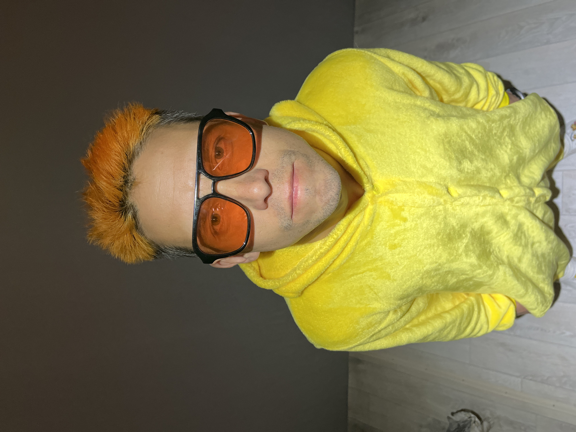 A man with short, orange hair and orange-tinted glasses is wearing a yellow hooded sweatshirt. The man is positioned indoors against a background with a gray wall on one side and wood-paneled flooring on the other. No information suggests this photo was taken in Chaikovsky, Russia.