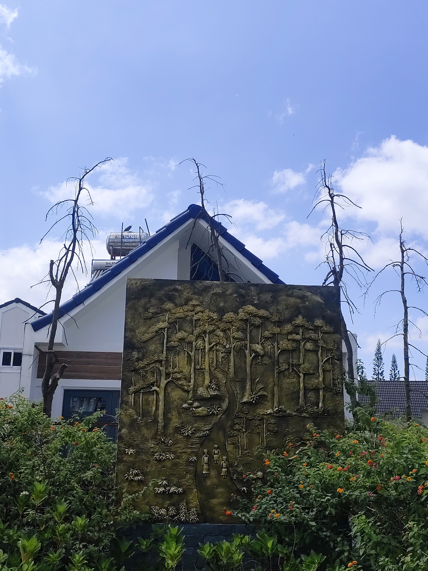 The photograph captures a scene in Lạc Dương, Vietnam, featuring a textured, rectangular monument depicting a forest scene with figures. The monument is positioned amidst lush green shrubbery with orange and white flowers. Behind the monument stands a two-story white building with a dark blue-tiled roof, topped with a solar water heater. Three bare trees are interspersed around the monument and building. The sky is light blue with scattered white clouds. Another building with a dark gray roof is partially visible on the right.