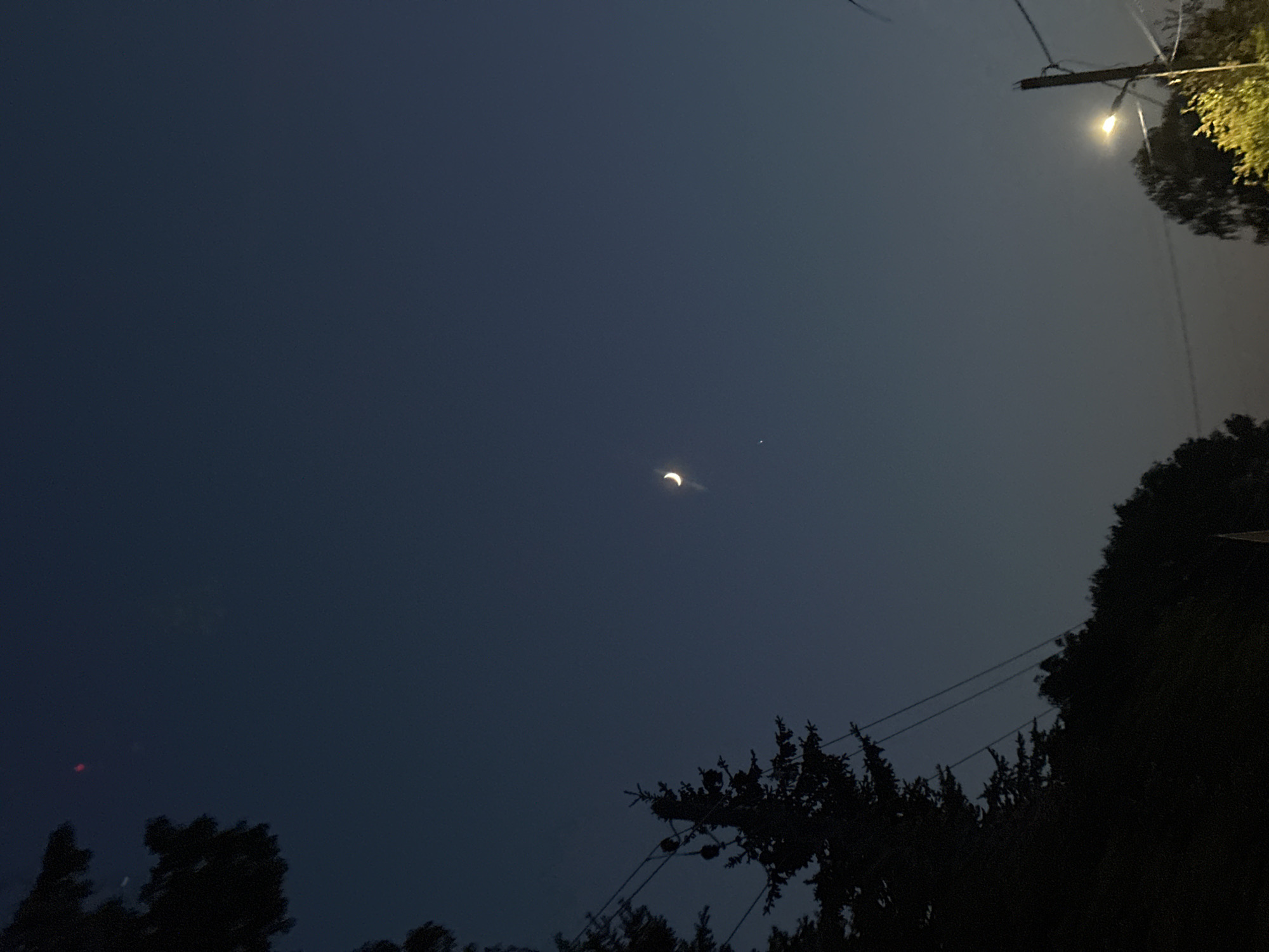 A night shot reveals a crescent moon in a dark blue sky, situated slightly left of center, with a faint dot possibly representing a star or planet appearing just to its right. Silhouetted trees are positioned in the foreground along the bottom, and to the right. Power lines stretch across the sky above these trees. A streetlight is visible at the top right corner, with some tree foliage near it. The photograph appears to be taken in an urban or suburban environment. Location information of Nizhny Novgorod, Russia, is irrelevant as the image does not reveal any clues about the location.