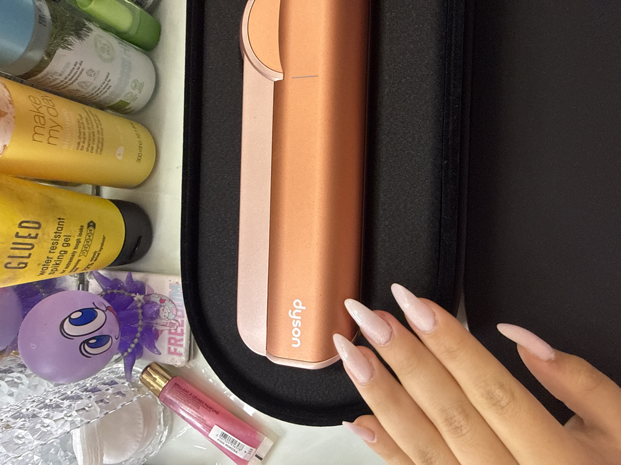 An overhead, slightly angled shot displays a collection of personal care products and a hand with manicured nails on a light-colored surface, possibly a vanity or countertop.
The primary subject is a rose gold or copper-colored Dyson hair styling tool, prominently displayed in a black case or holder on the right side of the frame. To its left, a hand with long, almond-shaped, light pink glittery fingernails rests near the Dyson product.
On the left side of the image, several items are visible. From top to bottom, partially obscured, is a white bottle with green accents, with text including "ANIC" and "DINAL WATO". Below it is a yellow bottle of "make my day shampoo," described as "milk shampoo for super soft hair" and containing "300ml e 10.1 fl oz". Another yellow bottle, labeled "GLUED water resistant Spiking gel," advertises "The extremely tough looks" and "12h wind Ingredients". Further down, a purple spherical object with large blue cartoon eyes is visible next to a white box labeled "FREZOOM". A small pink tube of "BRILLANT A LEVRES PARFUMÉ PINK MIMOSA" lip gloss lies nearby, along with crumpled silver foil and white cotton pads.
The scene suggests an indoor setting, likely a personal space for beauty and grooming. No specific activity is actively taking place beyond the arrangement of these items and the resting hand, implying a moment of preparation or display. The lighting appears to be bright and even, consistent with indoor illumination.