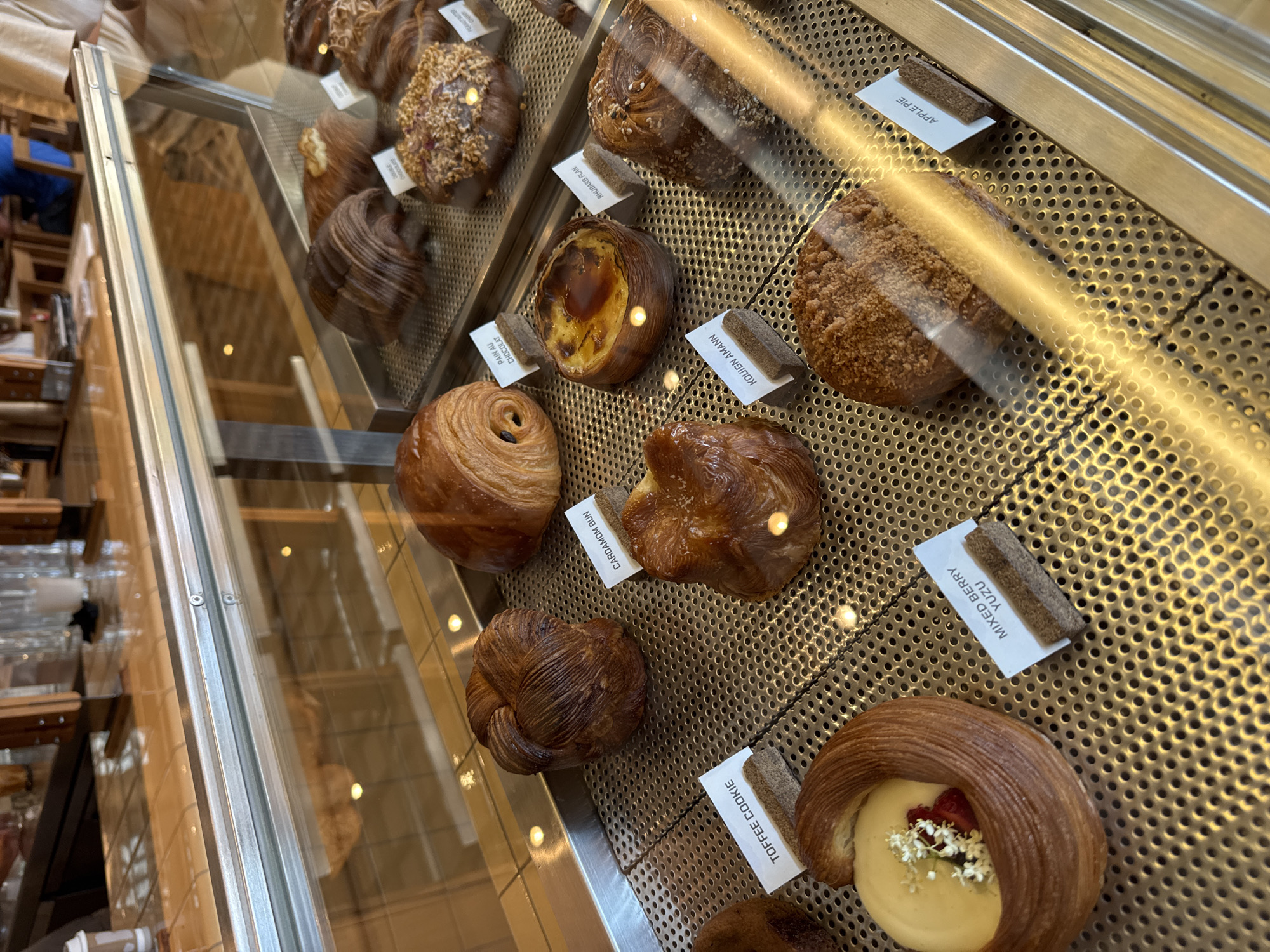 The image shows a display case in what appears to be a bakery or pastry shop. The case contains an assortment of baked goods, each with a small, rectangular tag identifying its name. From left to right and top to bottom, the pastries are: a croissant, a pastry covered in nuts, a similar looking pastry but with a different topping, a Pain au Chocolat, a Rhubarb Flan, a Kouign Amann, a Cardamom Bun, another pastry with a different shape, a Toffee Cookie, and a Mixed Berry Yuzu.
Based on the pastries and the general style, this image appears to be taken at the bakery "Bakesmith" in North Kuta, Indonesia.