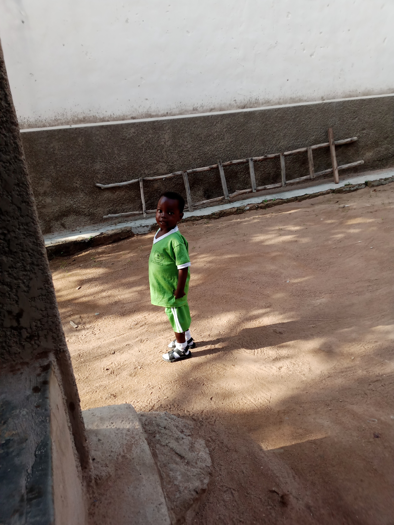 A young boy wearing a bright green t-shirt and matching shorts stands on a dirt ground in Jauro Yinu, Nigeria. The t-shirt has a white collar with green trim and a small emblem on the left chest. The shorts also have white trim along the sides. He is wearing white socks with black and white sandals. Behind him, a rough textured wall extends horizontally, with a lighter colored section above it. To the right of the wall, a rustic wooden ladder is propped up against it. The ground is covered in light brown dirt, with shadows from an unseen object casting dappled patterns across the scene. To the left of the boy and extending into the foreground, a rough, textured concrete structure, possibly stairs or a wall base, is visible. The overall environment appears to be an outdoor, somewhat unkempt area, possibly a courtyard or yard.