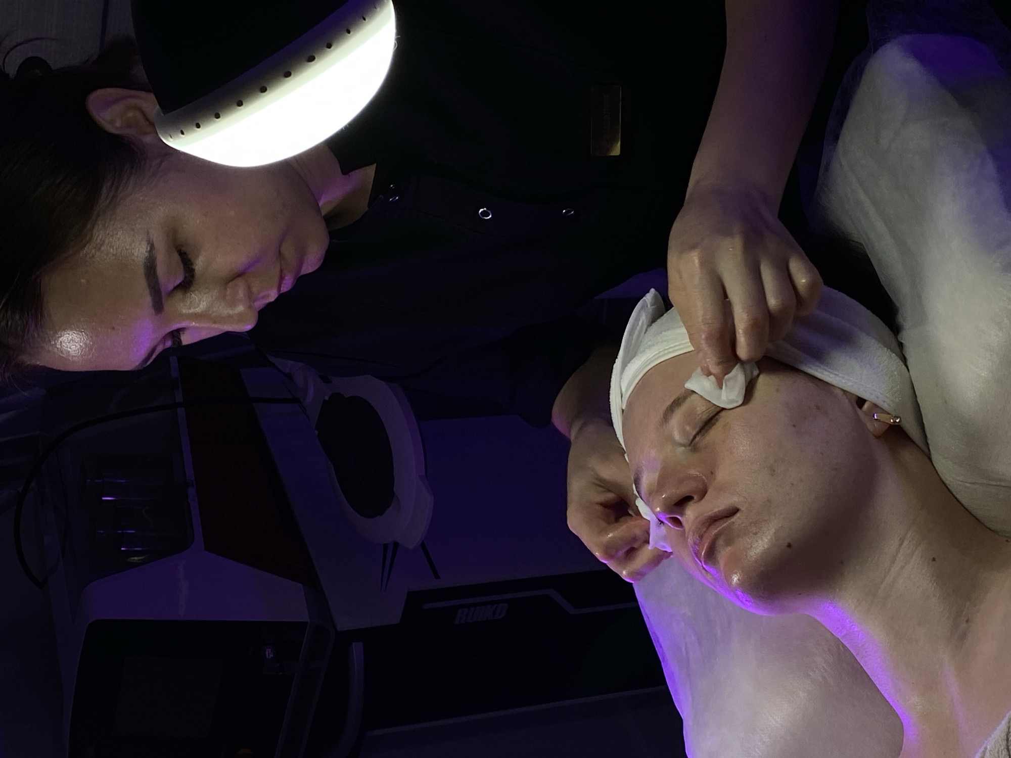 A medium shot depicts a woman lying on a white, possibly paper-covered surface, receiving a facial treatment. She has a white towel wrapped around her head and is wearing gold earrings. Her skin appears moist and has visible pores.
A person in a black shirt, presumably a beautician, is leaning over her, holding a white pad or cloth. Their hands appear wet, and they seem to be cleaning or applying something to the woman's face. A ring light is positioned close to the beautician's face.
The background is dark, but a piece of equipment with "RUKD" printed on it is visible on the lower left. The environment has a purple hue, possibly from lighting. The scene is most likely taking place in a beauty salon or spa. The location in the city of Izhevsk, Russia is not ascertainable from the visual data.