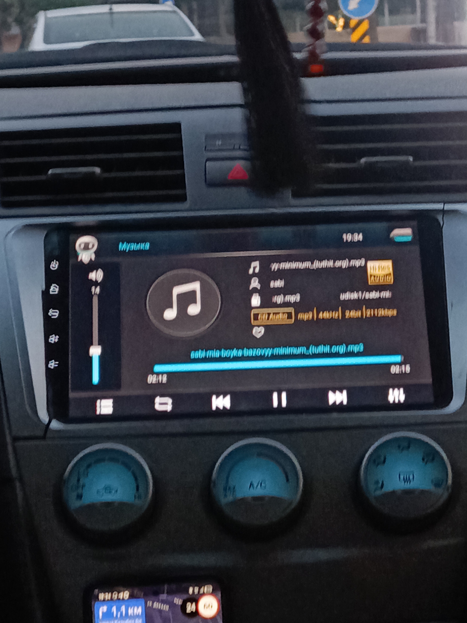 The image shows the interior of a car, looking towards the dashboard. A car stereo with a touchscreen display is the central focus. The display shows a music playback interface, with the song title "sabi mia boyka minimum__(tuthit.org).mp3" and playback controls. The time is displayed as 19:34. To the left of the stereo, there are several buttons and a volume slider indicating a volume level of 14. Below the stereo, there are three circular gauges; the left one shows "A/C". In the background, through the car's windshield, another car is visible. The scene appears to be in a city, likely Shymkent, Kazakhstan, given the context of the prompt. The overall lighting suggests it is either late afternoon or early evening.