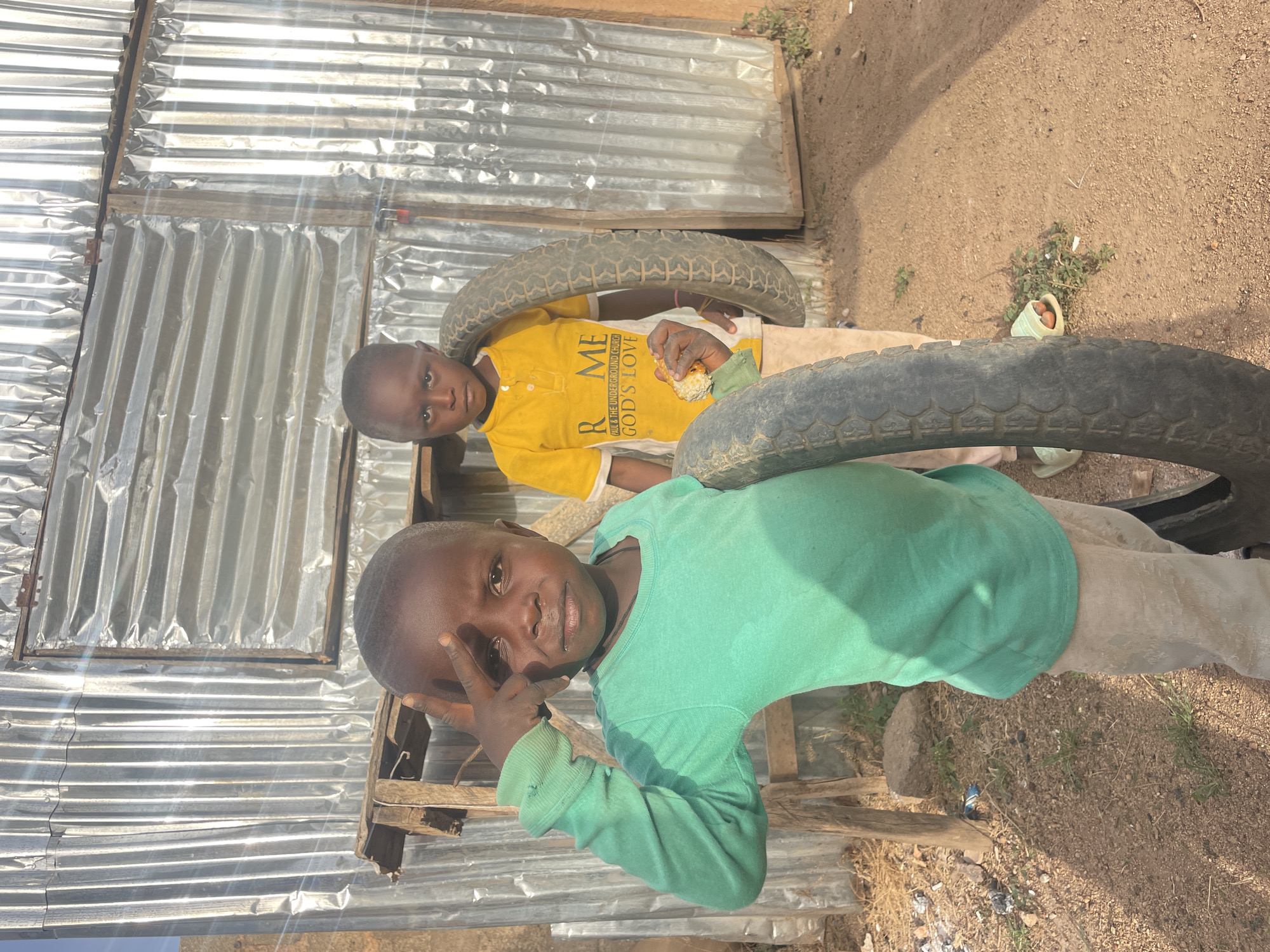 The image captures two young boys in an outdoor setting, likely in an informal settlement or rural area, consistent with Barakin Lerawi, Nigeria. The scene is brightly lit by direct sunlight, indicating daytime.
In the mid-ground, to the left, a young boy with dark skin and short hair is seen wearing a yellow short-sleeved shirt and light-colored pants. He is holding a piece of food, possibly corn or bread, in his right hand and appears to be eating. His shirt features visible text, which reads: "R ME / AND THE UNDERGROUND CHURCH / GODS LOVE". He is positioned near a large, dark tire.
Closer to the foreground, on the right, another young boy with similar features wears a long-sleeved, light green shirt and light-colored pants. He looks directly at the camera with a subtle smile, making a "peace" or "V" sign with the fingers of his right hand. He is also situated near another large, dark tire.
The background consists primarily of corrugated metal sheets, likely forming a wall or part of a dwelling, secured with wooden framing. The ground is dry, light brown dirt or sand, with sparse patches of green weeds and small rocks. The bright sunlight creates strong reflections and glare off the metal surfaces. The overall impression is one of children in a candid moment within a simple, rustic environment.