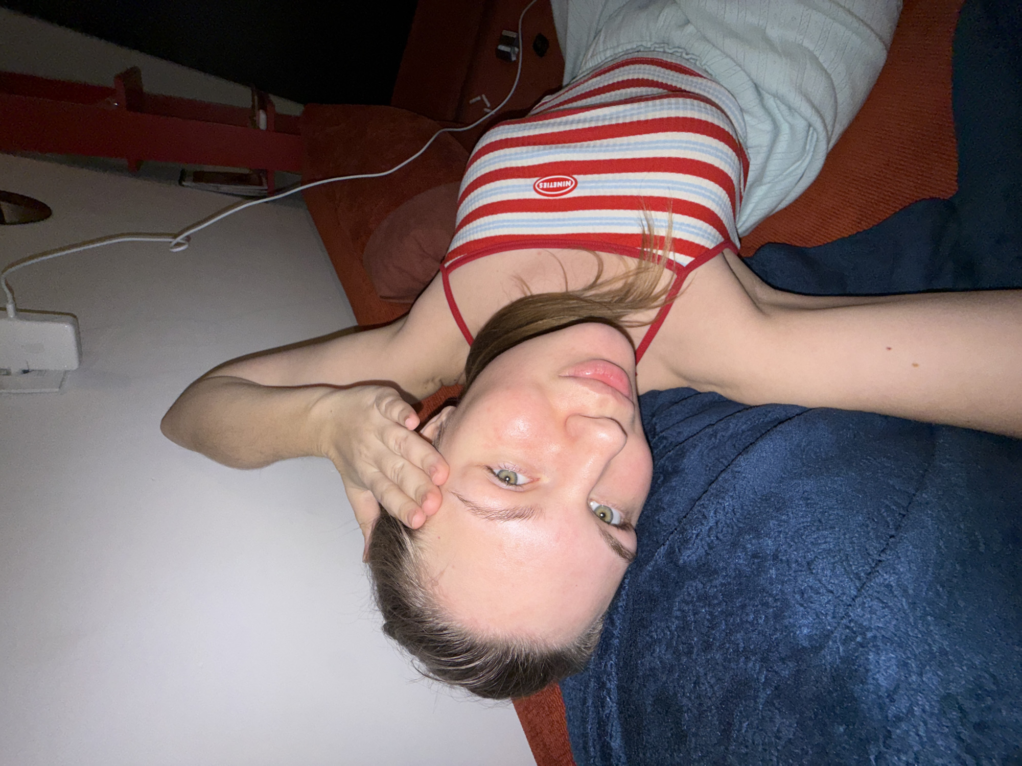 An image taken indoors in the city of Vozdovac Urban Municipality, Serbia, depicts a young adult female reclining on her left side. She has light skin, brown hair, and green eyes, and is looking directly at the camera with a slight smile. Her left arm is bent, with her hand resting on her forehead. She is wearing a red, white, and light blue striped tank top with thin red straps; a red oval patch on the top reads "NINETIES" in white text. Light blue, possibly ribbed, lower garments are also visible. Her head rests on a dark blue, textured cushion or blanket. To her right, an orange-red upholstered surface, likely a sofa or bed, extends across the background. A white charging cable hangs from a white electrical outlet or device mounted on a light-colored wall in the upper left. A red structural element, possibly part of furniture or shelving, is partially visible in the upper right background.