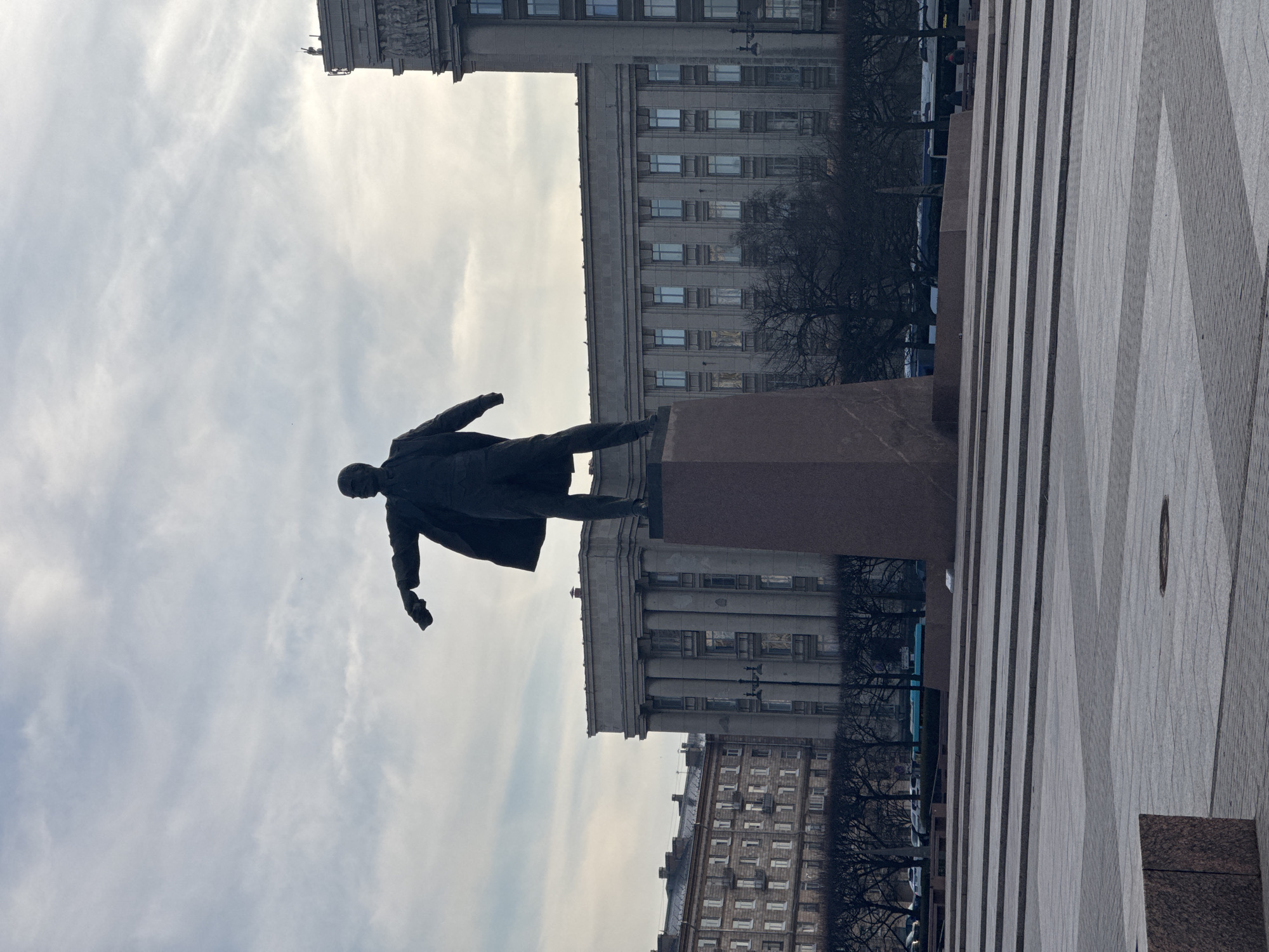 A statue of Vladimir Lenin stands atop a dark-red granite pedestal in front of a large, neoclassical building, likely a government or public building, in Saint Petersburg, Russia. The statue is made of a dark material, possibly bronze or iron. Lenin is depicted in a dynamic pose with his right arm extended. The square features a geometric pattern on the ground, with light-colored stone or concrete slabs. The sky is overcast, and there are bare trees in the background, suggesting it is autumn or winter. The overall scene conveys a sense of Soviet-era monumentality within a contemporary urban setting.