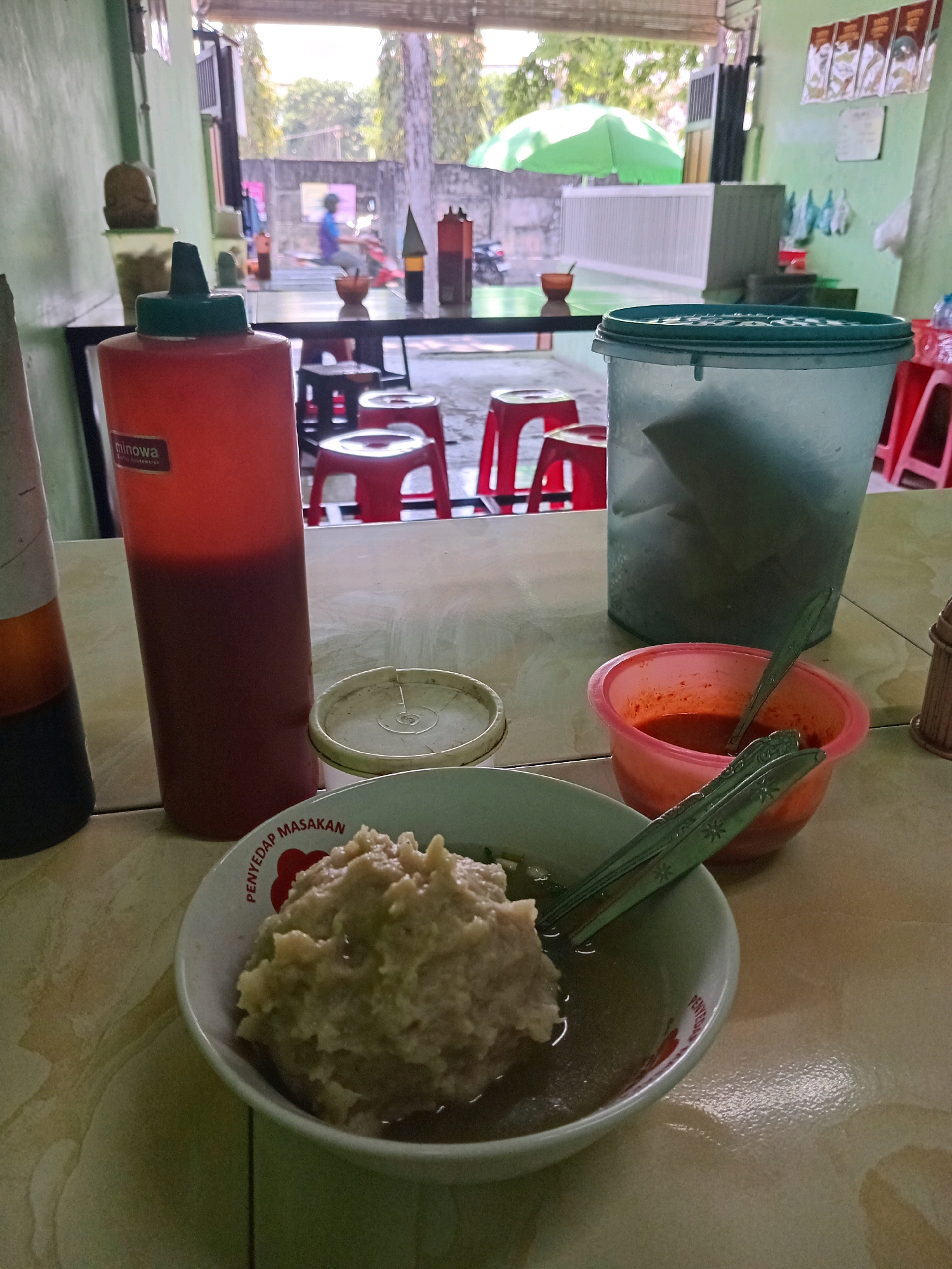 The image depicts a simple eatery in Tlogobendung, Indonesia. In the foreground, a bowl of bakso (Indonesian meatball soup) with a large meatball is prominently displayed, accompanied by a small bowl of chili sauce and a spoon. To the left, a large red squeeze bottle of sauce, likely chili sauce or ketchup, with a "Minowa" label is visible. Behind the food, a large, translucent blue plastic container, possibly for ice or drinks, sits on the table. Further back, several red plastic stools and a table suggest a casual dining setting. Through an open doorway in the background, trees and a street scene with a person on a motorbike can be seen, indicating the eatery is open to the outdoors. The walls are painted a light green.