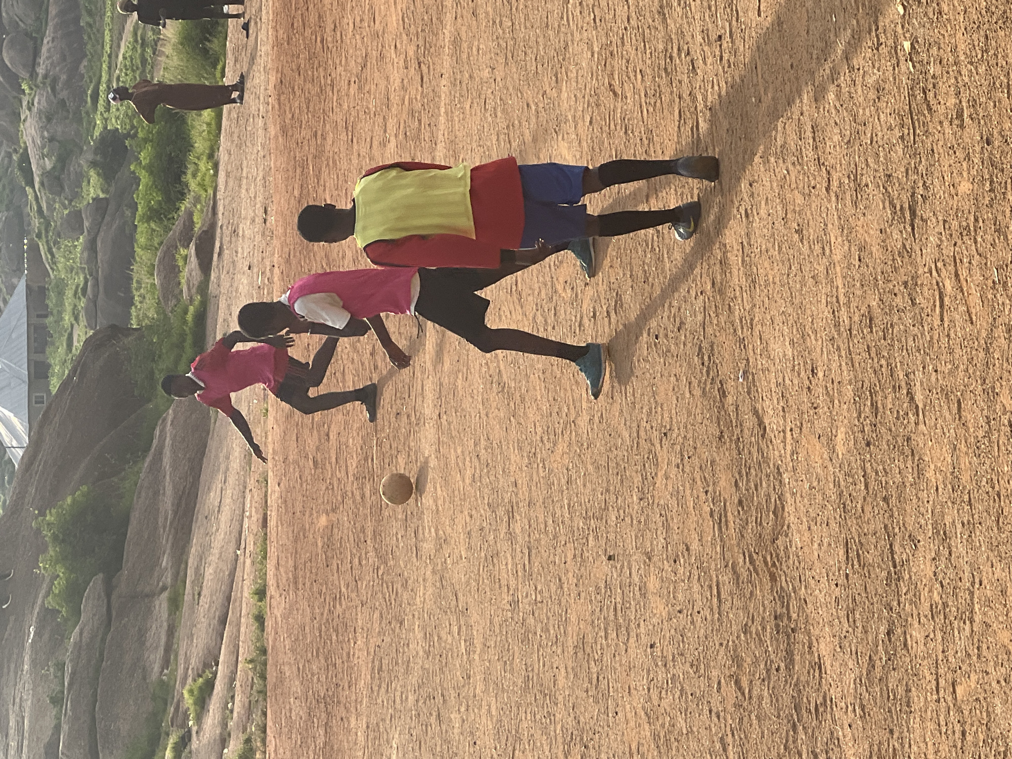 In the city of Jos, Nigeria, a group of young men are playing soccer on a dirt field. The field is dry and dusty, with rocks and sparse vegetation in the background. The players are wearing casual clothing and some have donned bibs. One player in a pink shirt is in the process of kicking a soccer ball, while another in a yellow bib is positioned to receive a pass. Two other players are visible further in the background, observing the game. The scene appears to be set in the late afternoon, as evidenced by the long shadows cast by the players. The overall atmosphere is one of casual recreation and community engagement in a rugged, natural setting.