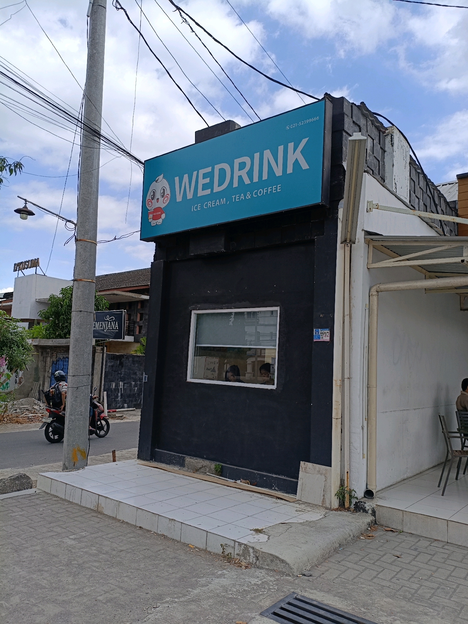 The image depicts a street-level view of a commercial establishment named "WEDRINK" in Maguwoharjo, Indonesia. The "WEDRINK" sign is a bright turquoise rectangle with white lettering and a cartoon mascot. Below it, the text reads "ICE CREAM, TEA & COFFEE". The building itself is partially dark, with a contrasting white section to the right, featuring an outdoor seating area with chairs. A utility pole with numerous wires runs vertically along the left side of the frame. In the background, other buildings are visible, including one with a sign that says "D'KOSMA". The street in front of the establishment shows a paved road and sidewalk area. A person on a motorcycle is in motion on the road. The sky is partly cloudy.