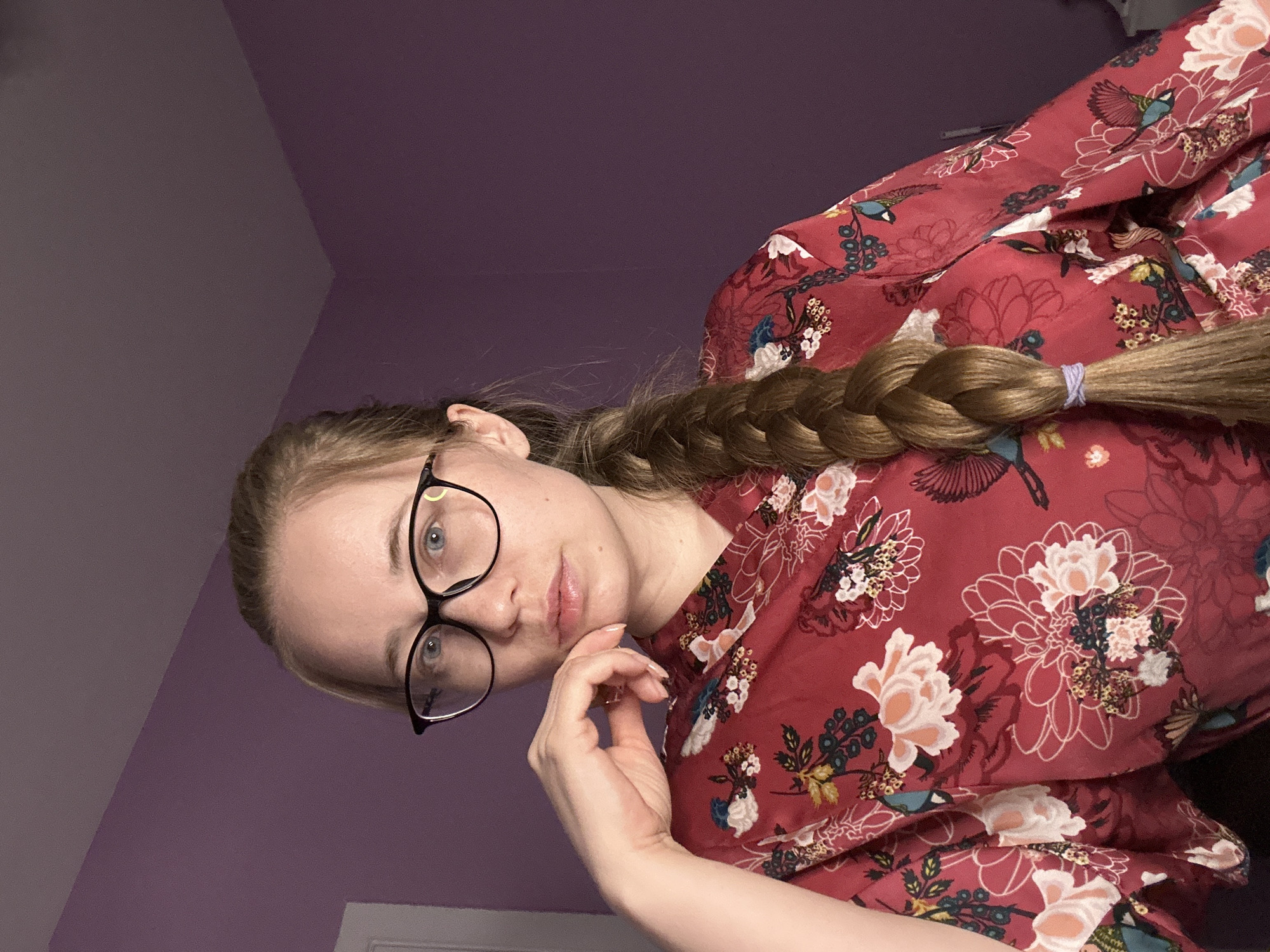 A woman is pictured indoors against a purple wall with a gradient color change. She wears black-rimmed glasses and a red floral blouse. Her hair is blonde and styled in a braid. Her right hand is raised, with her fingers gently touching her face. There is no evidence to suggest this image was taken in Saint Petersburg, Russia.