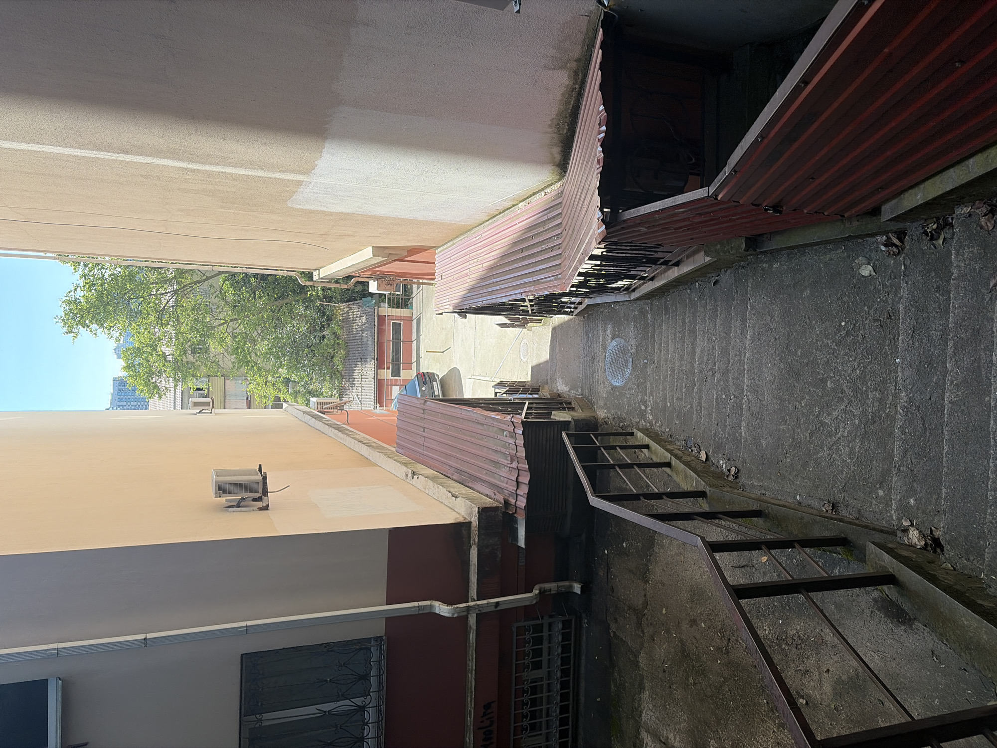 This media file captures a narrow, downward-sloping outdoor passageway or alleyway in an urban setting, likely in Sochi, Russia, as suggested by the context. The scene is brightly lit by direct sunlight under a clear blue sky, indicating daytime.
The passageway is bordered by buildings on both sides. On the left, a multi-story building features light beige-colored stucco walls. An external air conditioning unit is mounted on its upper section, and a lower portion of the wall is painted a dark red/maroon, with windows protected by intricate metal grates. A white gutter system runs down this side. Above, the lush green foliage of trees and a bright blue sky are visible, with hints of taller buildings in the distance.
On the right, another building is partially obscured by a reddish-brown corrugated metal awning or roof that extends over the passageway, showing signs of wear and moss. Below this, a concrete staircase with metal railings descends into a darker, shadowed area. The concrete steps are worn and scattered with dry leaves. Further down the path, circular drain covers are visible on the ground, and a small car is partially seen in the distance.
The strong sunlight creates stark contrasts between illuminated and shadowed areas, emphasizing the texture of the building walls and the worn concrete surfaces. No people are present, and no specific event or activity is taking place. Graffiti spelling "TOOLITA" is visible on the lower dark red wall on the left side of the passageway. The overall impression is that of an older, functional urban corridor.
