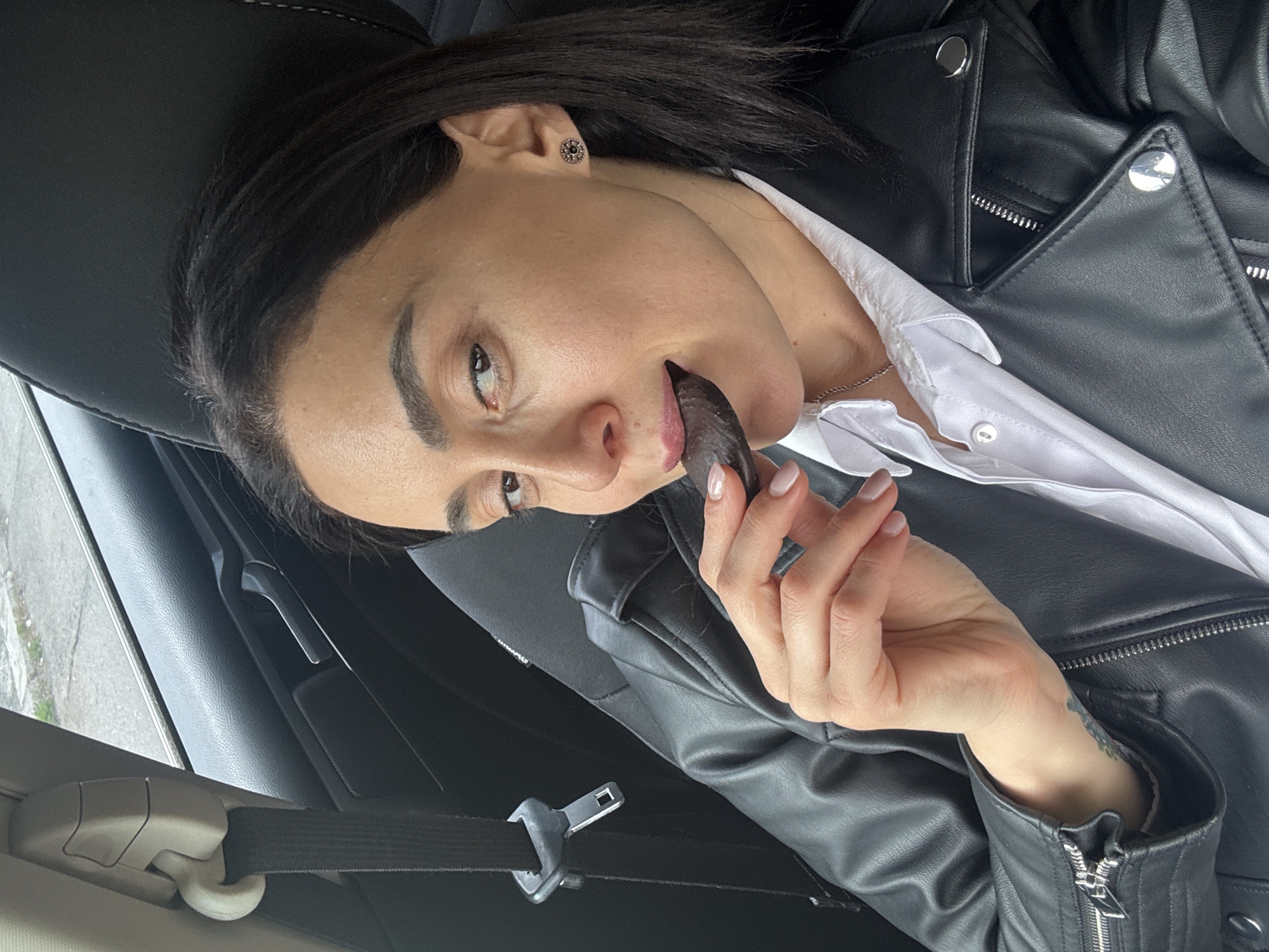 A woman with dark hair is sitting in the driver's seat of a car, wearing a black leather jacket over a white collared shirt. Her seatbelt is fastened. She is holding a dark, cylindrical object to her mouth. The interior of the car is visible, with the window and door trim partially shown. The location is likely Tbilisi, Georgia.