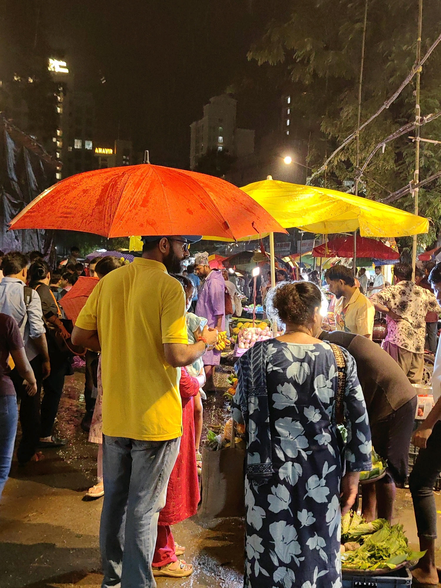 This is a nocturnal street scene in Mumbai, India, possibly a market area. The image is dominated by several colorful umbrellas, a large red one in the foreground and a bright yellow one behind it. People are gathered under and around these umbrellas, suggesting they are sheltering from rain or browsing goods. The ground appears wet, indicating recent or ongoing precipitation.
In the foreground, a man wearing a yellow t-shirt and blue jeans stands facing away from the camera, holding a red umbrella overhead. To his right, a woman in a dark blue, floral-patterned dress is visible from behind. She is looking at a stall laden with green leafy vegetables in a black crate.
In the midground and background, more people are milling about. Some individuals are dressed in traditional Indian attire, such as the man in a purple garment to the right. Others are in casual Western clothing. There are glimpses of stalls with various items, including what appear to be fruits and vegetables. The lighting is dim, with some artificial lights illuminating the scene, and the background shows buildings with illuminated windows, characteristic of an urban setting. The overall atmosphere is that of a bustling, wet, night market.
