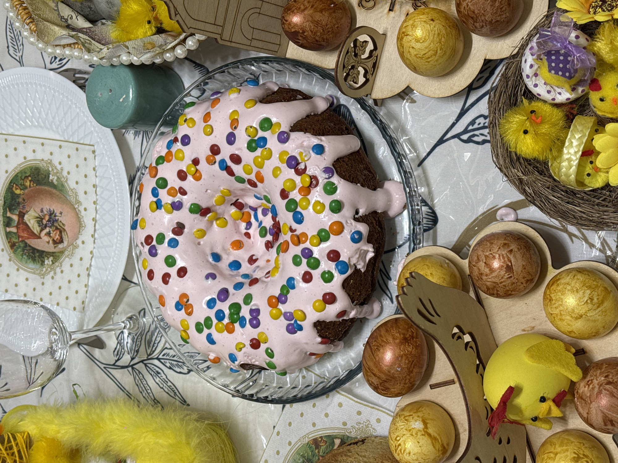 The image shows an Easter spread, likely in Yerevan, Armenia. A ring cake is the focal point, decorated with pink frosting and colorful candy sprinkles. Various Easter decorations surround the cake, including dyed eggs in gold and brown hues, decorative chicks, a candle, a decorative plate, and a wine glass. The tablecloth is patterned with leaves, adding a festive touch to the scene.
