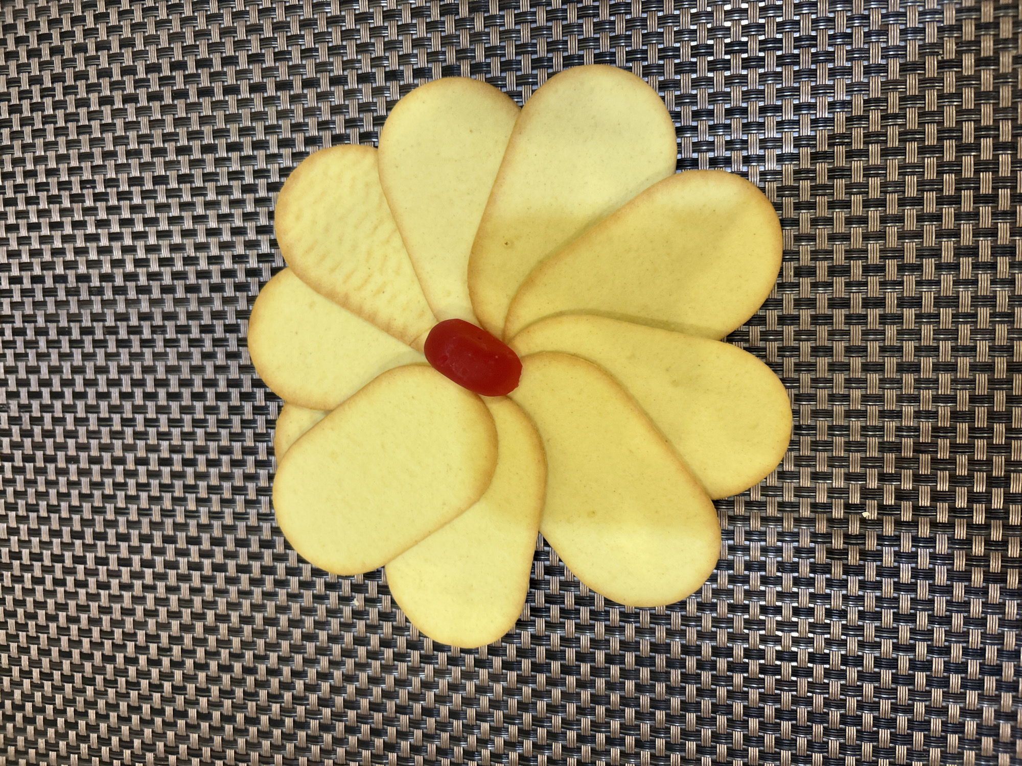 A close-up, top-down shot features a flower-like arrangement of light yellow oval cookies on a dark brown and tan woven placemat. A single, bright red piece of candy sits at the center where the cookies meet. The cookies are arranged in a circular pattern, overlapping each other to form petal shapes. The lighting is bright and even, creating soft shadows and highlighting the texture of the placemat and the surface of the cookies. No people or specific city locations are visible in the image.