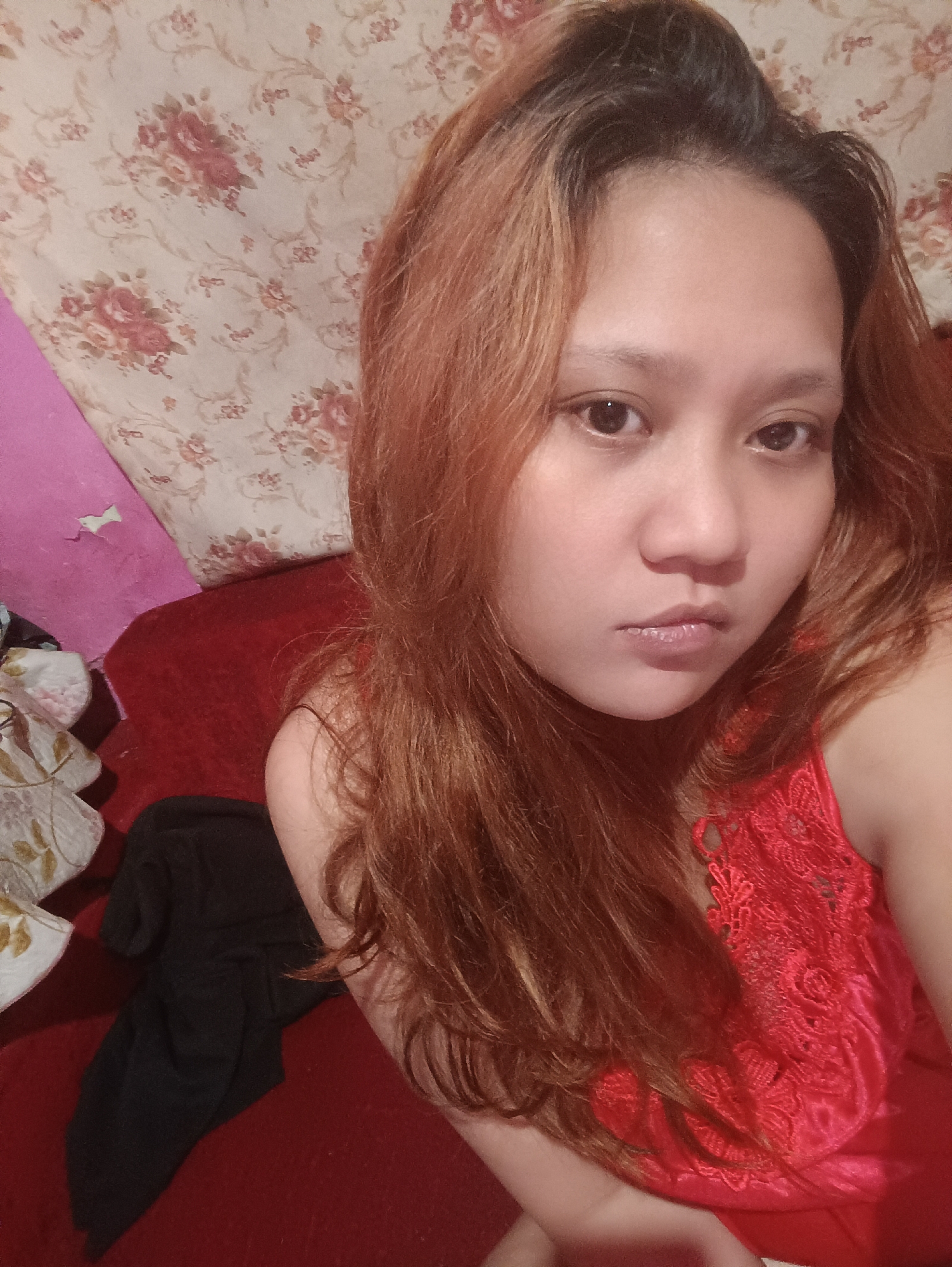 A young woman with long, wavy, reddish-brown hair is captured in a close-up selfie. She is wearing a red lace-trimmed garment. Her face is turned slightly towards the camera, with her gaze directed forward. Her expression appears neutral. The background consists of a red upholstered surface, likely a sofa, with a wall behind it adorned with a floral wallpaper pattern featuring pink and cream roses. A folded black garment rests on the red surface to her left. The lighting is somewhat dim, suggesting an indoor setting. The scene is likely a private residence in Depok, Indonesia, given the mention of the location.