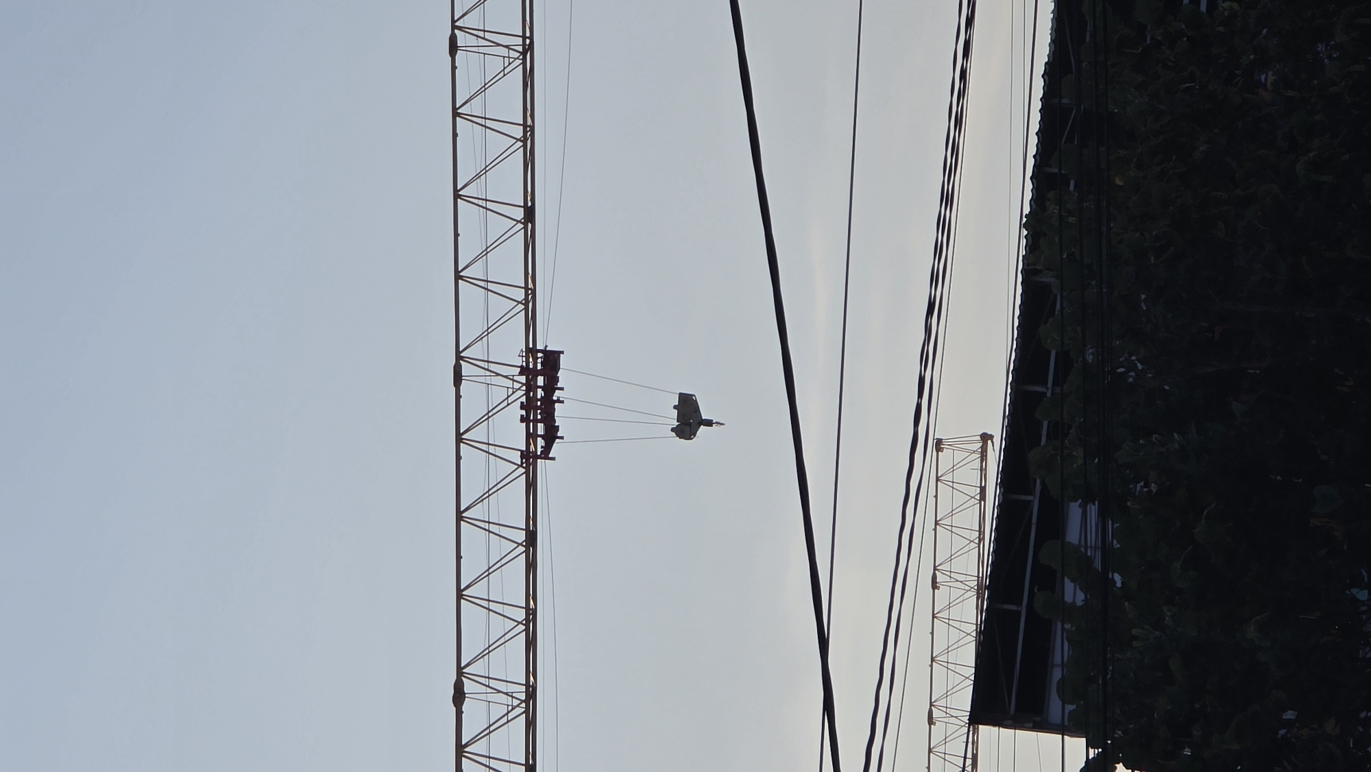 The image, presented in a portrait orientation (rotated 90 degrees clockwise from its natural horizontal perspective), depicts an outdoor scene dominated by industrial infrastructure and a clear sky, set in Nha Trang Ward, Vietnam. A large, reddish-brown lattice structure, characteristic of a crane or tall tower, extends vertically along the left side of the frame. From this structure, a grey, rectangular trolley or hook block is suspended by multiple dark cables.
Several dark, thin lines, likely power lines, traverse the middle section of the image, running diagonally. On the far right, a dark, angled structure, possibly the roofline of a building, is visible, partially obscured by a dense mass of dark green foliage. Another smaller, lighter-colored lattice-like structure can be seen in the bottom right corner. The sky is a uniform light blue, suggesting clear daytime conditions, with a subtle gradient indicating early morning or late afternoon light. No people are visible, and no specific activity is taking place. There is no visible text in the image.