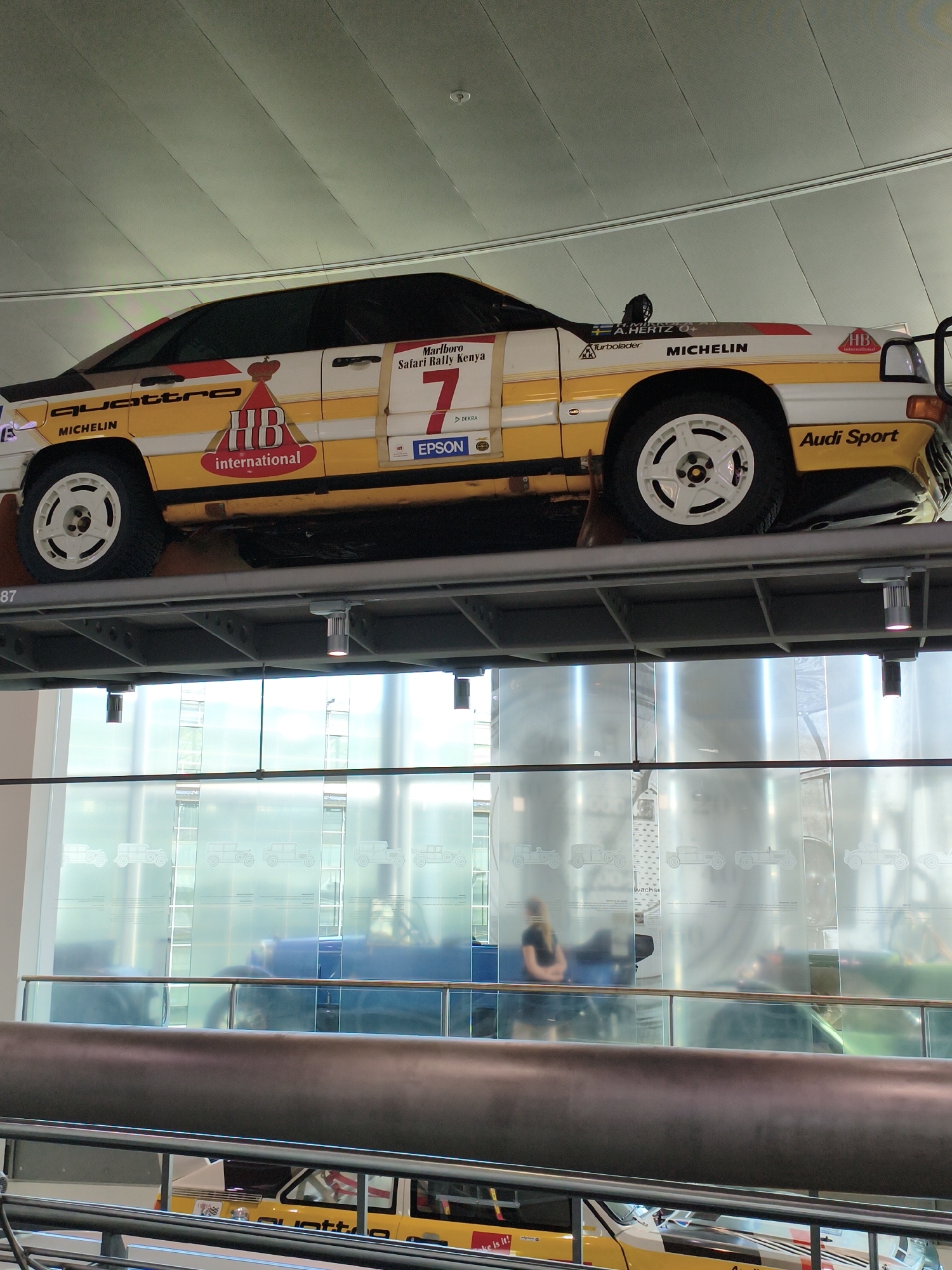 An Audi Quattro rally car, painted white and yellow with red accents and various sponsor logos, including "Marlboro Safari Rally Kenya," "HB International," "quattro," "MICHELIN," "EPSON," and "Audi Sport," is displayed on a metal platform. The car is seen from a slightly elevated side-front angle. Below the platform, a glass partition features etched silhouettes of vintage cars and a blurred reflection of a woman. The environment appears to be an indoor exhibition space, possibly a museum or showroom, with a high ceiling and bright lighting. The image is taken in Ingolstadt, Germany, at the Audi Museum mobile.