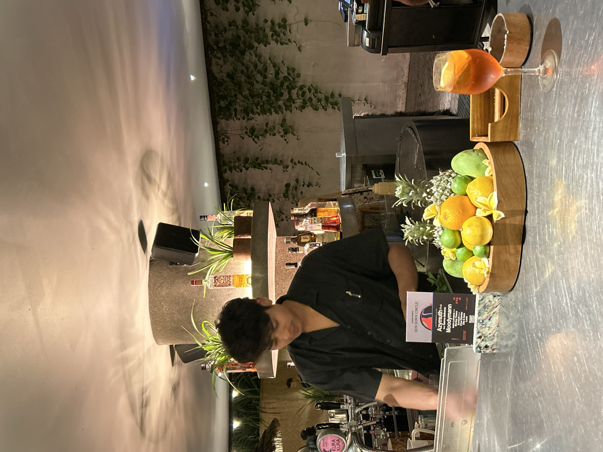 A young man in a black short-sleeved shirt stands behind a bar in what appears to be a dimly lit establishment in North Kuta, Indonesia. The bar is made of stainless steel and is covered in a light sheen. In front of the man is a wooden bowl filled with various fruits, including oranges, limes, and what appears to be a pineapple and mango. A cocktail glass containing an amber-colored drink with a slice of lemon is placed to the right of the fruit bowl, with an ashtray next to it. The background shows shelves stocked with bottles of alcohol, plants, and decorative vines hanging from a textured wall. The ceiling features overhead lighting with shadows cast from ceiling fans. A sign on the bar indicates an event called "SUN DOWN CIRCLE" featuring "Azimuth (live)" and "Moodymann". The man is in the process of preparing or serving something, with his hands near a clear plastic container.