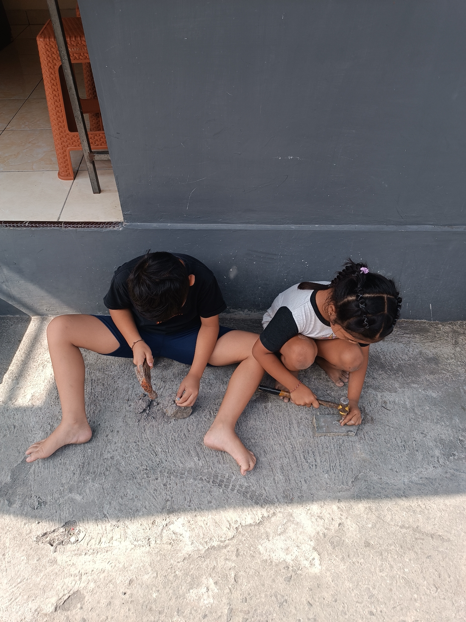 Two children, a boy and a girl, are sitting on a concrete floor in what appears to be an outdoor area. The boy, on the left, is wearing a black t-shirt and blue shorts. He is holding a piece of metal or stone and what looks like a hammer or similar tool. He is looking down at the object in his hands. The girl, on the right, is wearing a white t-shirt with black sleeves and dark shorts. She is kneeling and holding a small hammer and appears to be working on a small metal object on the ground. Her hair is styled in braids with a small purple accessory. Behind them is a dark gray wall. To the far left of the image, partially visible, is an orange plastic chair and some tiled flooring. The scene is lit by sunlight, casting shadows on the ground. This setting is consistent with a residential area in Tabanan, Indonesia, possibly a patio or yard.