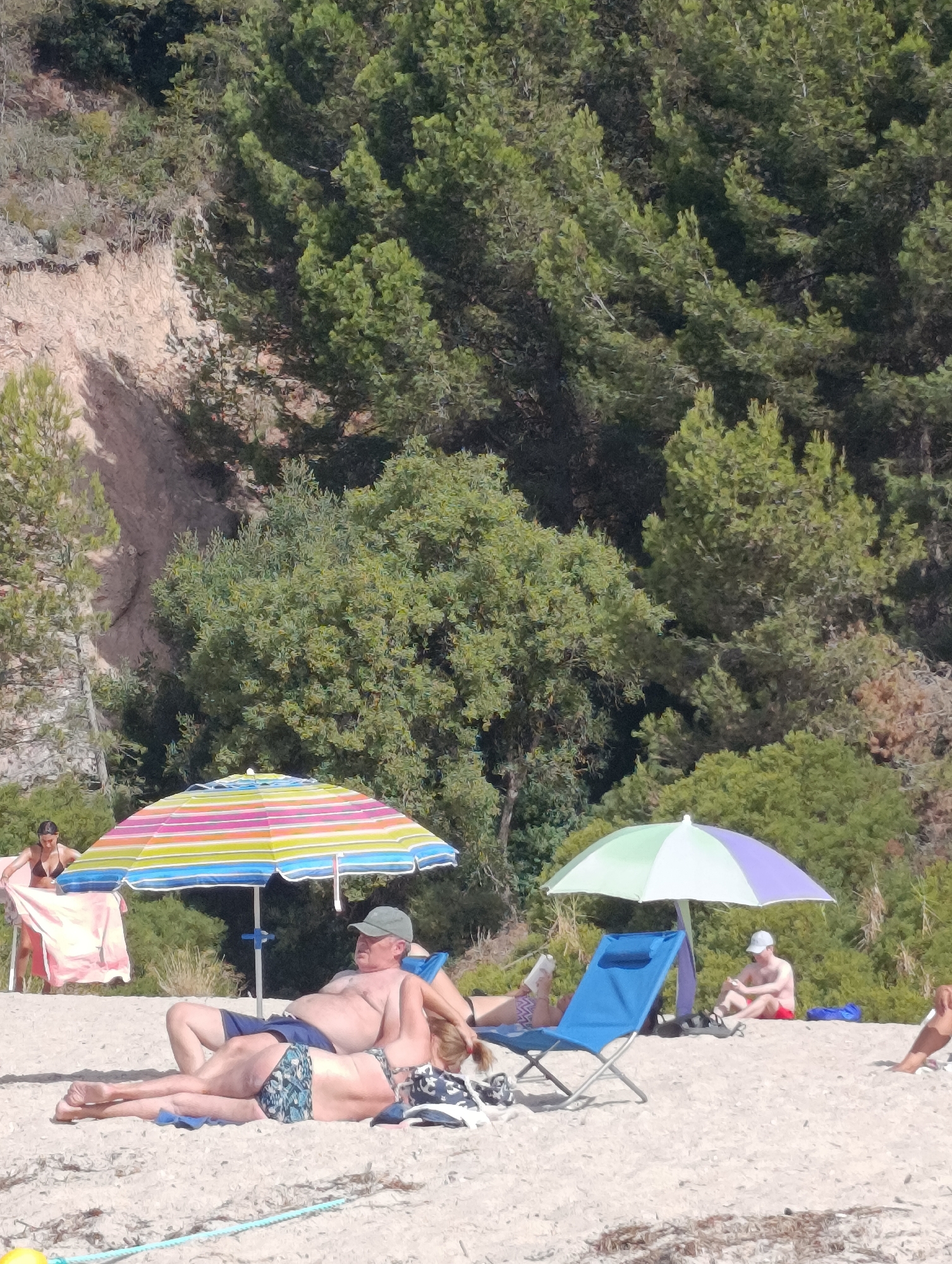 The image depicts a sunny day at a sandy beach, likely in Setúbal, Portugal, judging by the vegetation and coastal appearance. In the foreground, a couple is relaxing on the sand. The male is lying on his back, wearing blue shorts and a green cap, while the female is lying on her side, facing away from the camera, wearing a patterned bikini. They are positioned near a large, multi-colored beach umbrella. To the right, another umbrella, this one purple and green, shades a blue beach chair where a man in red shorts is sitting and looking at his phone. In the background, a woman in a bikini is standing near a pink towel. The beach is backed by a densely vegetated hillside, with trees and rocky outcrops visible. The sand is light brown and appears to be a typical beach environment with some scattered dried vegetation.