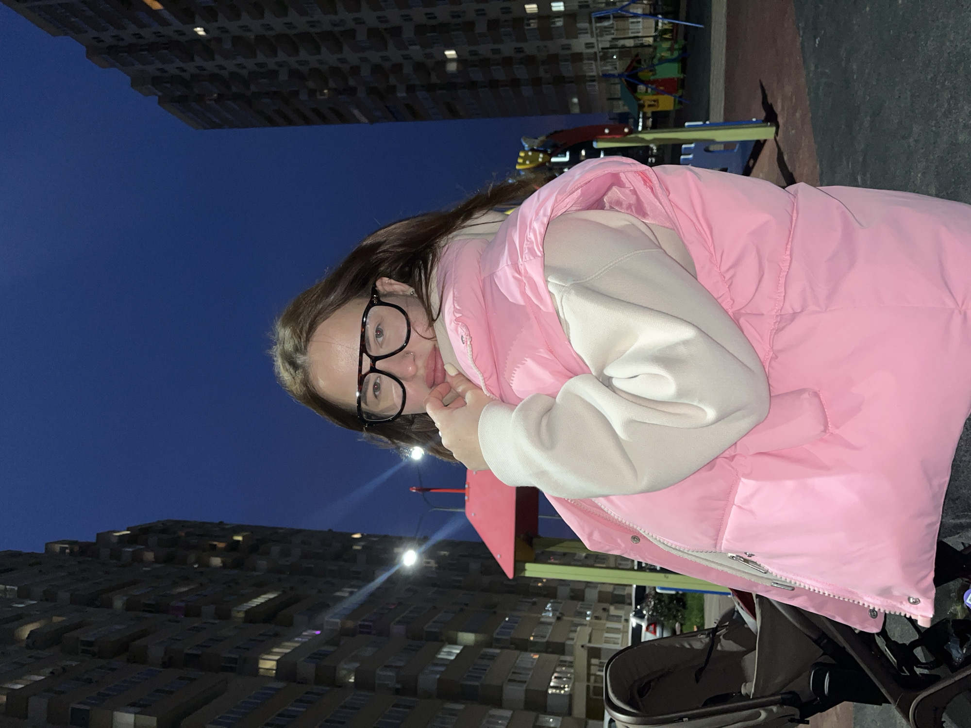 The image, rotated 90 degrees clockwise, depicts a young person outdoors in the evening. The main subject is a individual with long, dark hair and black-framed glasses, looking directly at the camera with a subtle smile. They are wearing a light beige or off-white hooded sweatshirt layered under a bright pink puffer vest. One hand is gently placed near their mouth or chin.
In the background, a deep blue twilight sky dominates, indicating it is evening or night. Several large, multi-story buildings, likely residential, are visible on both sides of the frame, with numerous windows illuminated, casting a glow. To the right of the subject, hints of a colorful playground structure can be seen, suggesting a recreational area. In the lower right foreground, a portion of a baby stroller is visible, implying the presence of a child or that the person is pushing it.
The overall setting is an urban residential area at night, possibly a courtyard or park within a city, with the cool weather suggested by the puffer vest. There is no visible text in the image. Given the provided context, the scene is located in Krasnodar, Russia.