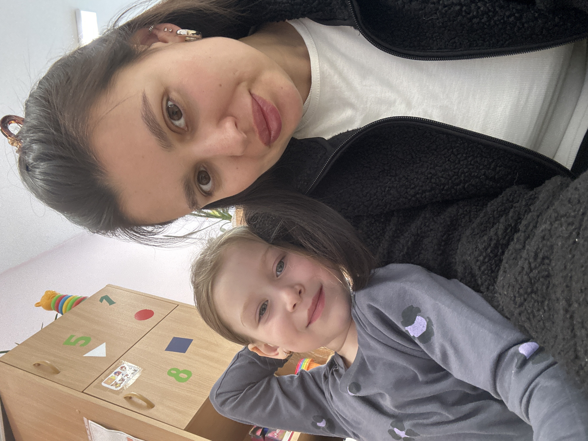 A close-up, tilted portrait captures an adult female and a young girl smiling at the camera in an indoor setting in Chaikovsky, Russia.
The adult female, with dark hair and brown eyes, is positioned at the top of the frame. She wears a black fuzzy jacket over a white t-shirt. Her lips are tinted, and her left ear features multiple piercings, including small hoops and a decorative cuff on the helix. She has a gentle smile.
Below her and slightly to the right is a young girl with light brown hair and blue-gray eyes, smiling broadly with her head resting on her right arm. She is wearing a grey long-sleeved shirt adorned with purple and dark grey animal print patterns.
In the background, behind the young girl, is a light-colored wooden storage cabinet or unit. The cabinet doors feature colorful educational stickers, including a green "1", a red circle, a green "5", a white triangle, a blue square, and a green "8". A small, clear sticker or card with various colorful shapes is also visible on one of the cabinet doors. On top of the cabinet, a vibrant, segmented toy, possibly a caterpillar, adds a pop of color. The lighting suggests it is daytime, creating a bright and relaxed atmosphere. Both individuals appear happy and comfortable.