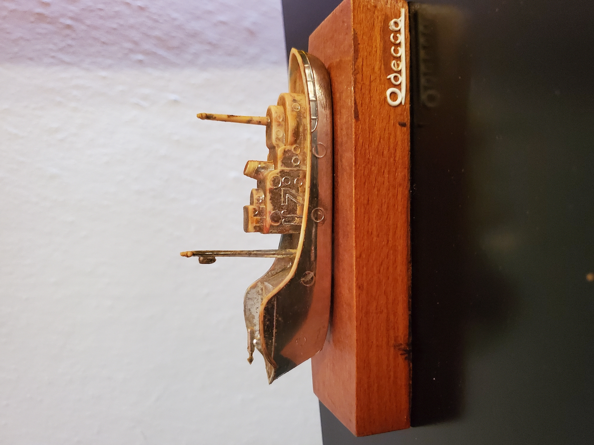 A close-up, eye-level, medium shot shows a small, metallic toy boat mounted on a rectangular wooden base. The boat is detailed with structures resembling cabins and a smokestack. The base is made of polished wood and has the name "Odecca" inscribed in white lettering on its right side. The scene is set against a textured, off-white wall. The object appears to be on a dark, reflective surface, possibly a table. The image does not provide any information about the city of Konstantinovka, Moldova, nor does it contain any people or actions.