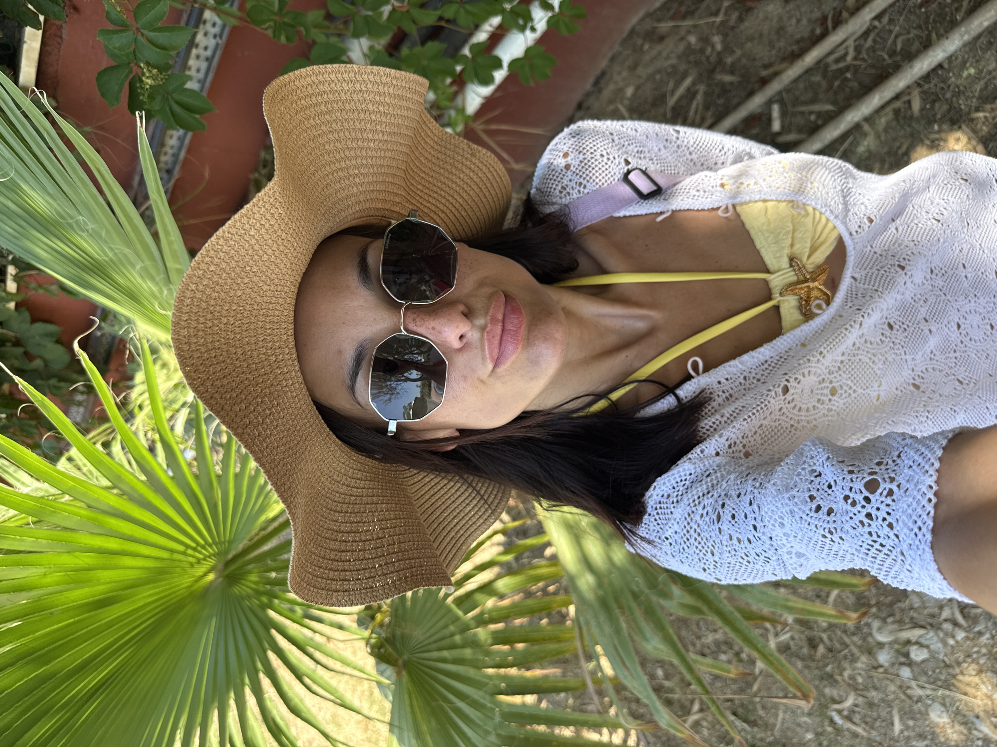 In an outdoor setting, a woman stands wearing a wide-brimmed straw hat, sunglasses, a yellow bikini top with a starfish pendant, and a white crochet cover-up. She is positioned in front of a large green palm frond, with greenery and a reddish wall visible in the background. The ground appears to be dirt with some scattered pebbles and fallen leaves. The lighting suggests it is daytime, possibly with strong sunlight. The location is identified as Unknown, Türkiye.